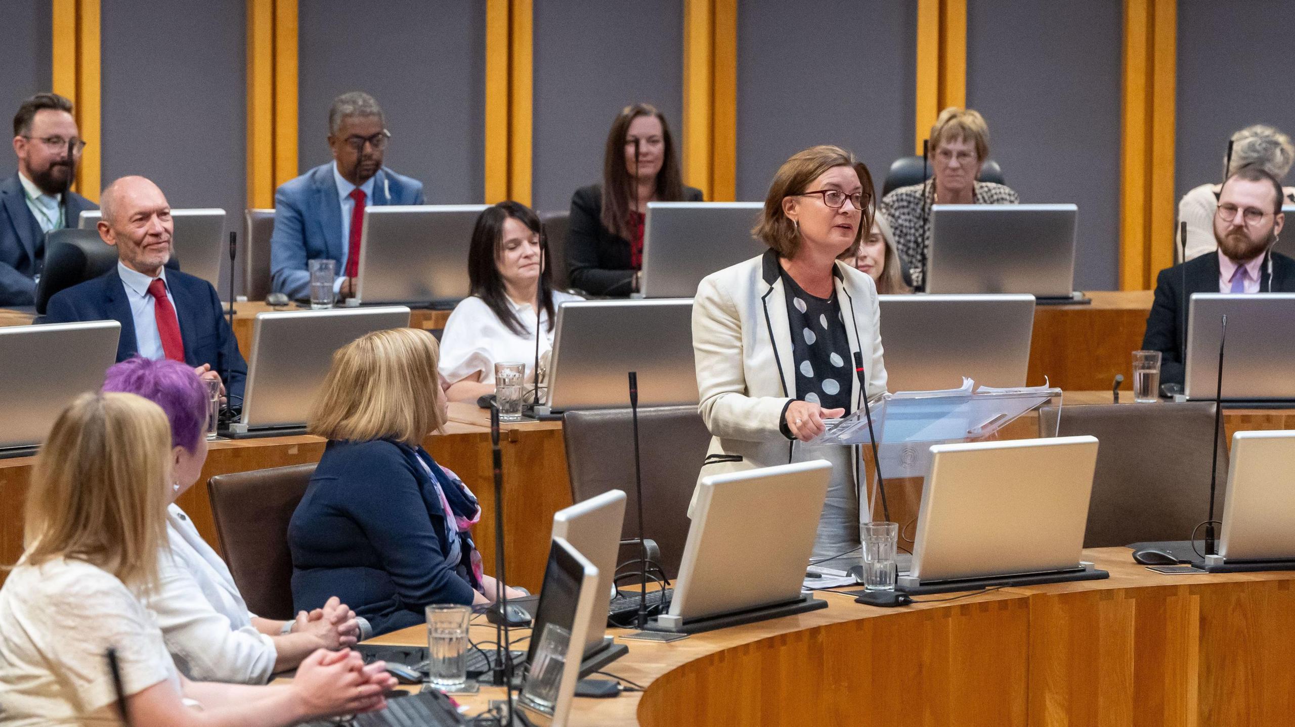 Eluned Morgan wearing a white jacket and standing, addresses the Senedd with other Labour politicians listening 