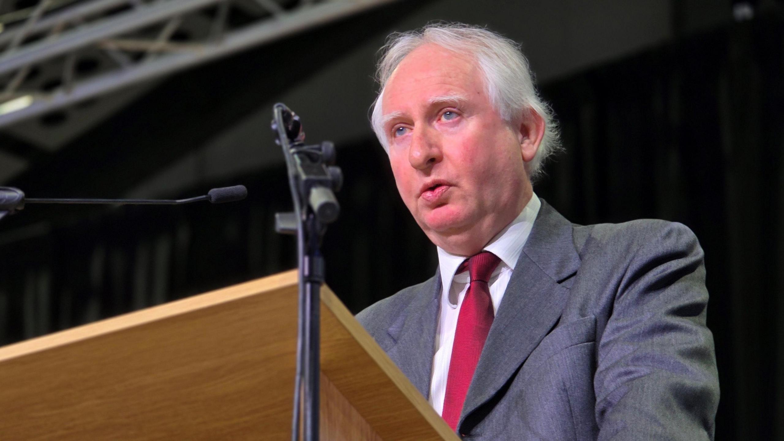 Daniel Zeichner is standing at a conference podium. He has a grey suit jacket on top of a white shirt and red tie.