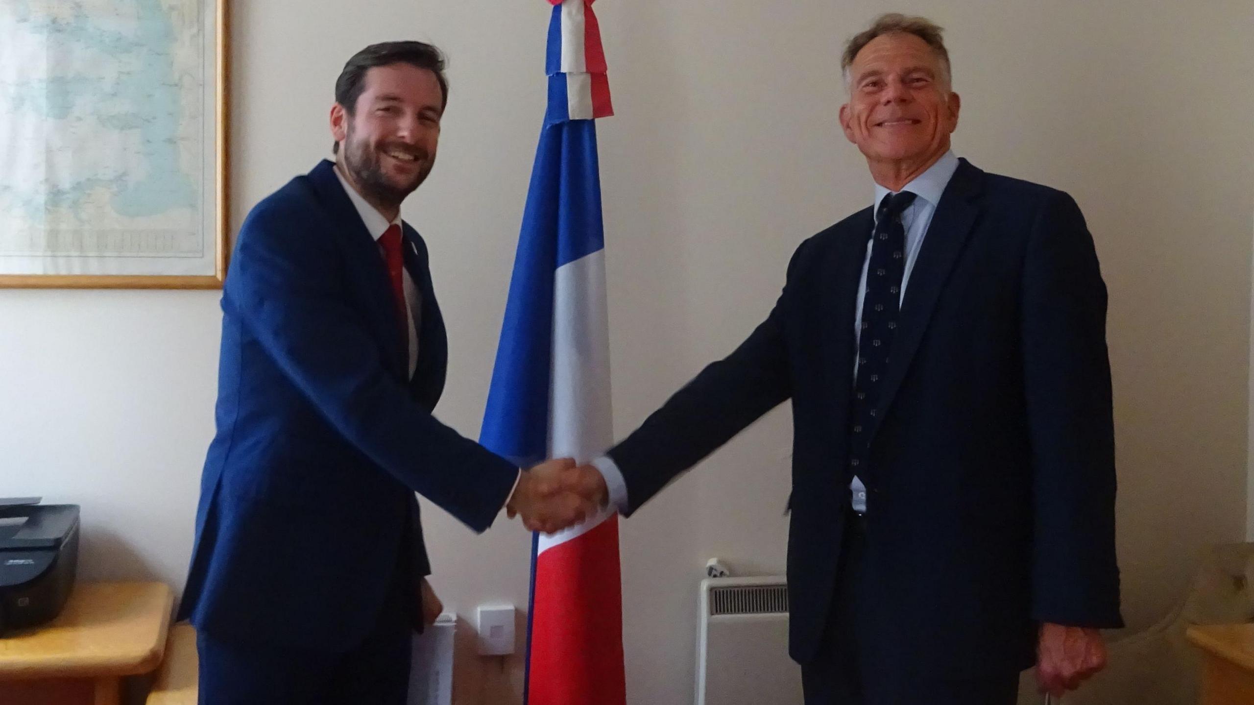 Pierre Chays and Christopher Scholefield in suits shaking hands in front of a French flag in an office