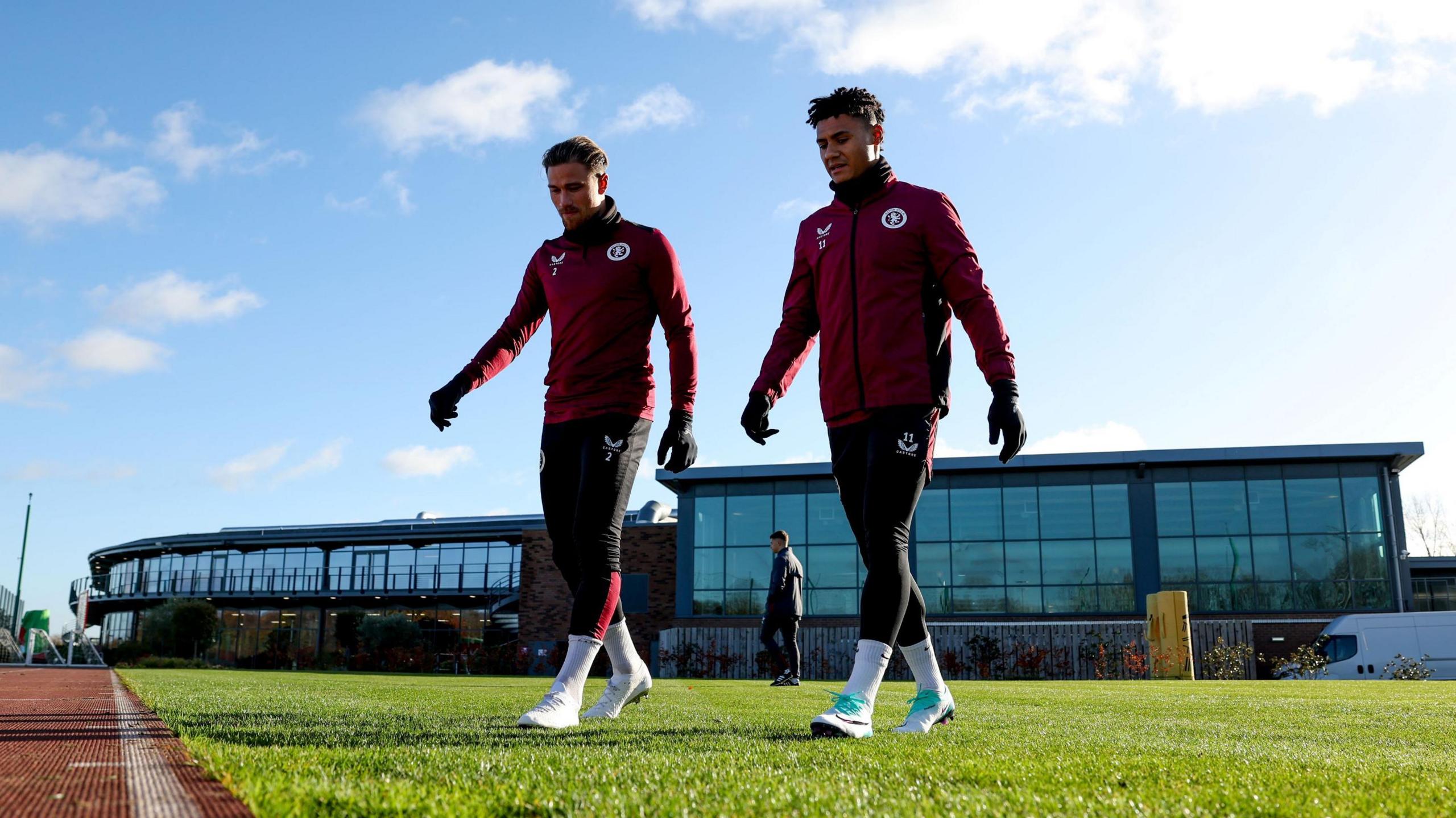Matty Cash and Ollie Watkins in maroon Aston Villa training kit walking in front of the glass-fronted Bodymoor Heath training ground