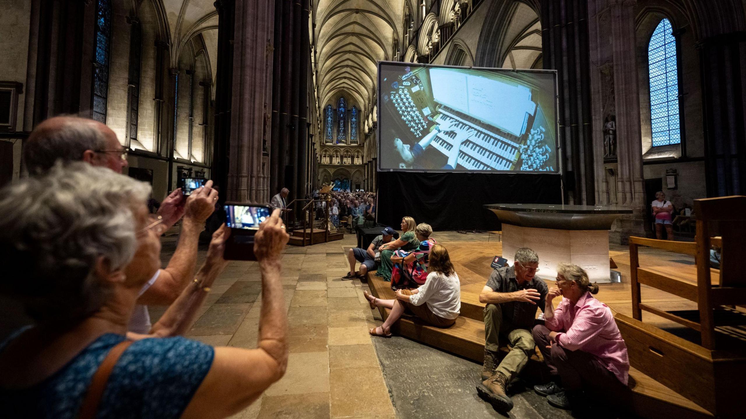 A large screen shows a man playing a cathedral organ in Salisbury, his hands of the keys and sheets of music. People sit in the large cathedral behind it, enjoying the organ music, with one woman taking a picture on her phone