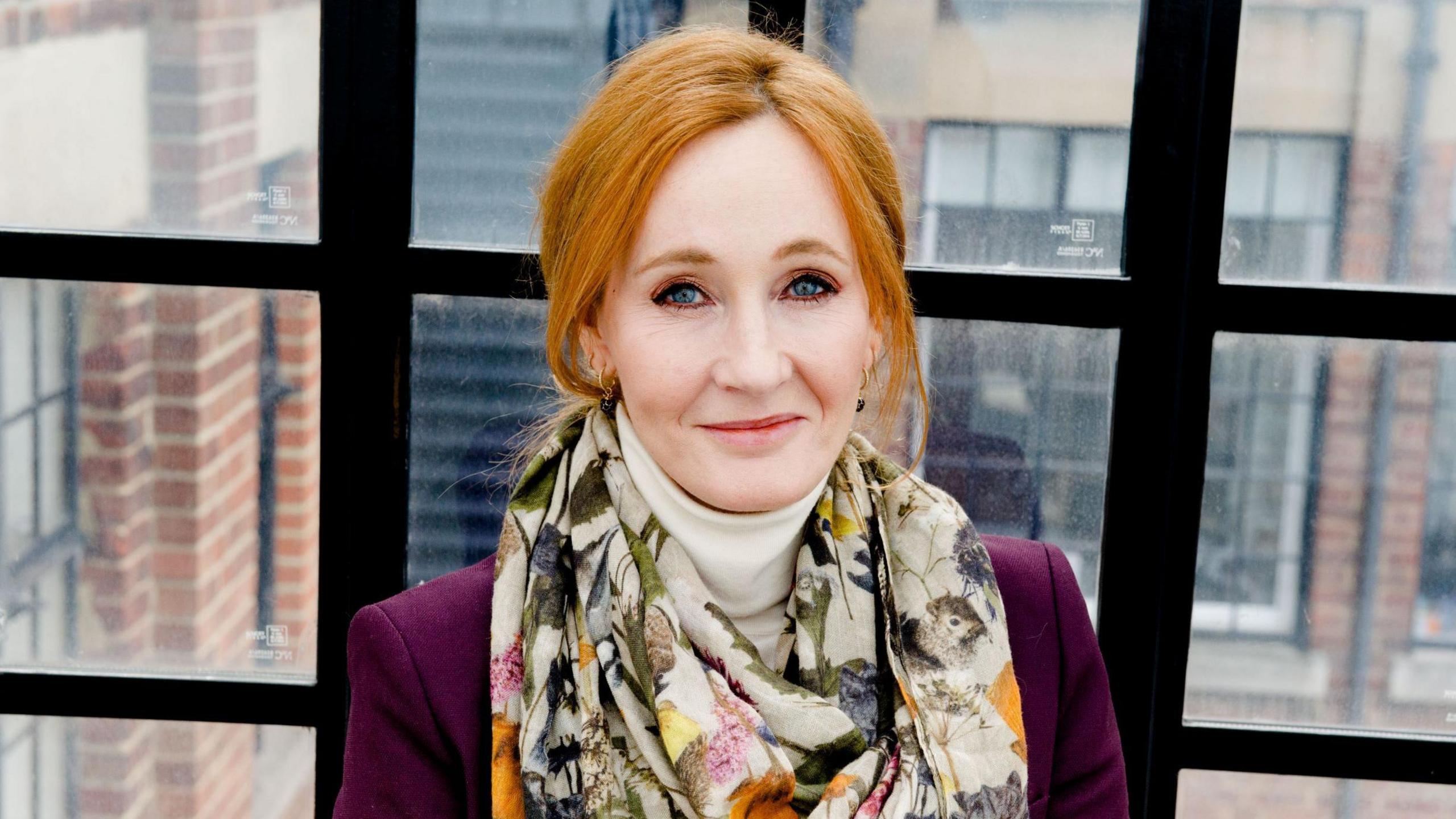 The author JK Rowling, sitting  by a glass window and looking at the camera. She has a multi-coloured scarf wrapped around her neck.