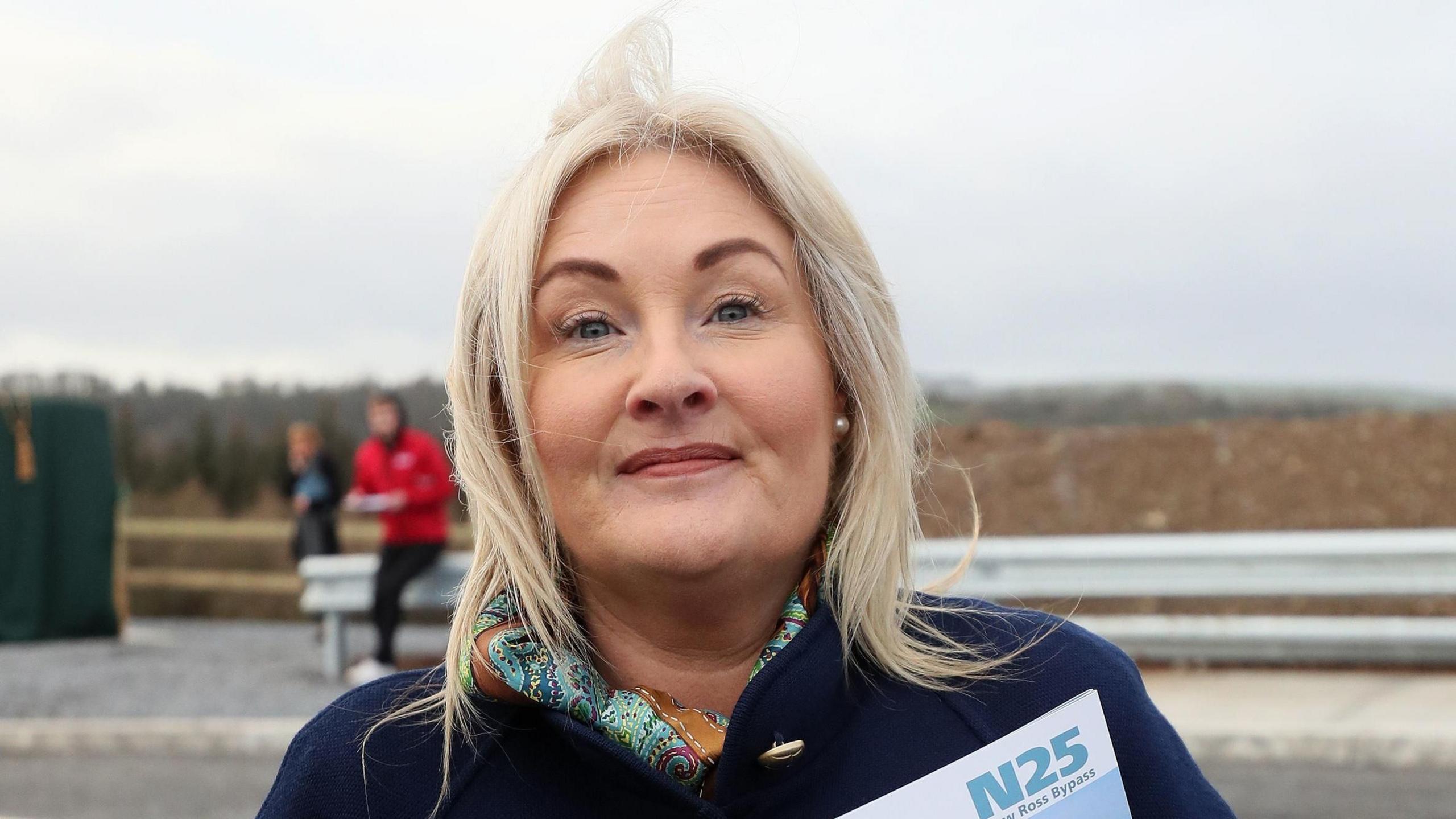 Verona Murphy wearing her blonde hair down with a navy coat on standing in front of a coast line 