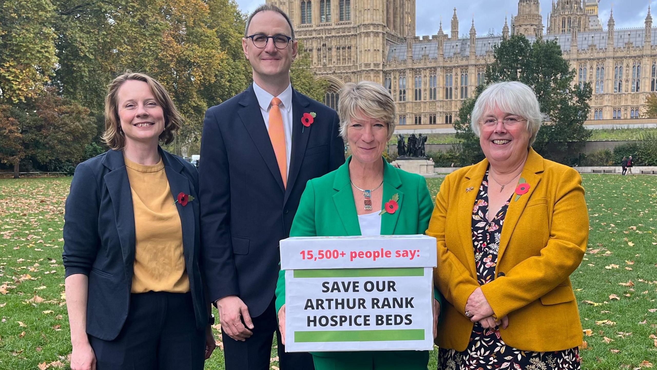 A group of four people standing outside. Pippa Heylings who is standing third from the left is dressing in a green suit and is holding a white box which says, 15,500+ people say: SAVE OUR ARTHUR RANK HOSPICE BEDS.