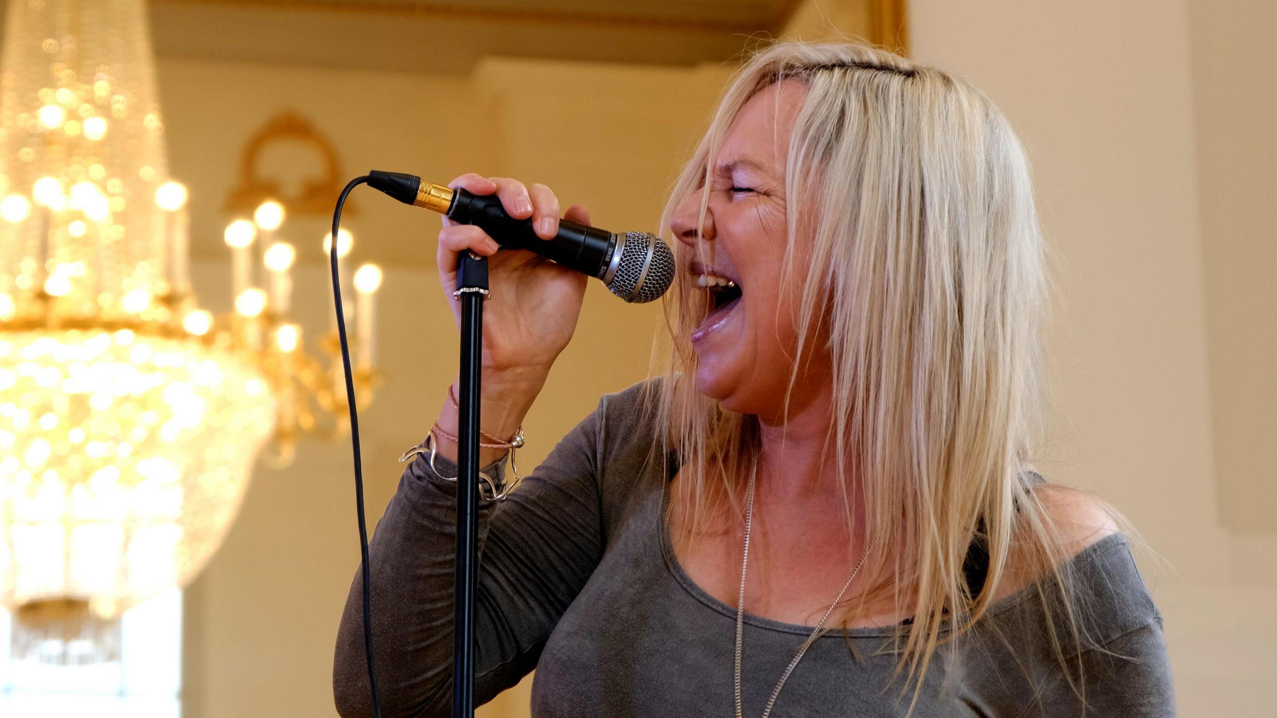 A woman wearing a grey long sleeved top and with long blonde hair singing passionately into a microphone in a room with a golden chandelier