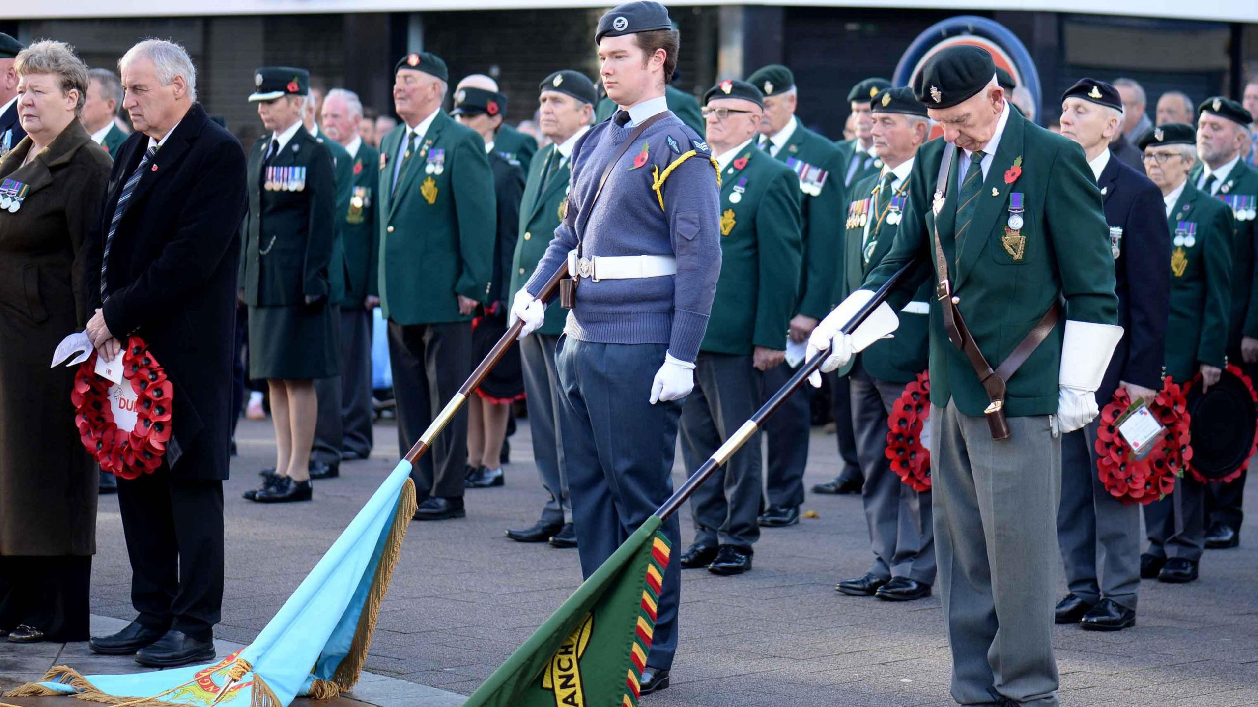 Two men are holding flags. Some men are holding poppy wreaths. A crowd of people behind them are dressed in uniforms. 