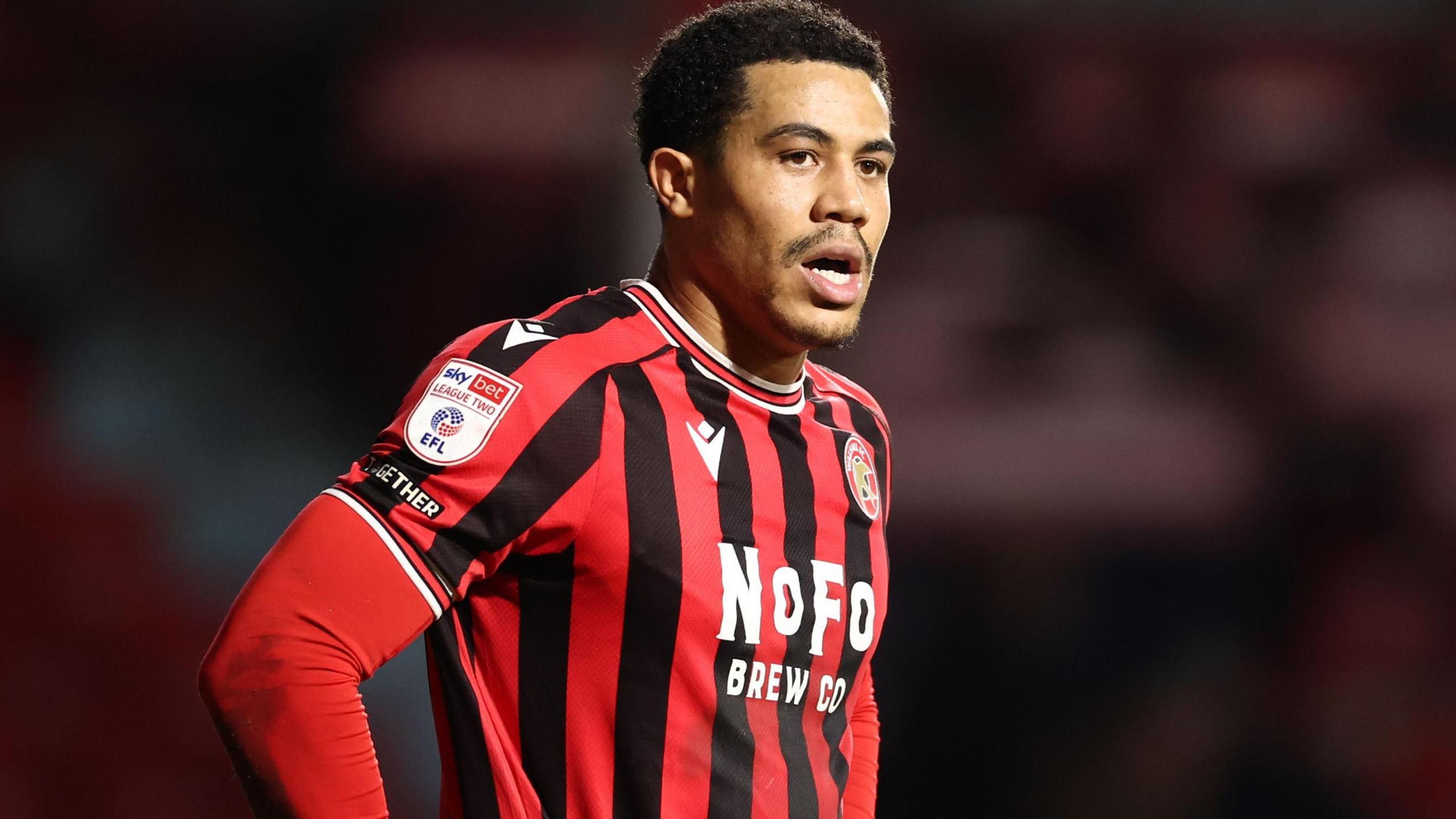 Priestley Farquharson in red-and-black striped Walsall shirt and red undershirt looks on during a game