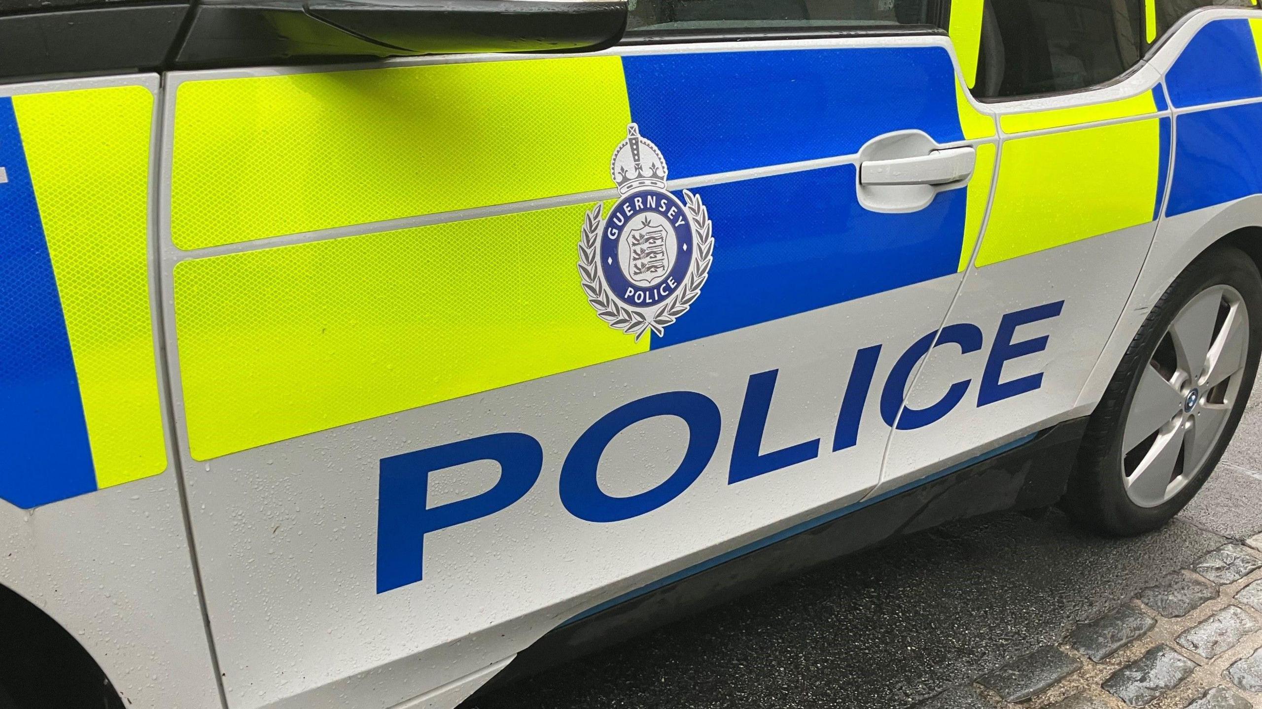 A picture of a police car parked on a road in Guernsey. The car is blue and green in colour and has the word POLICE in big writing on the side of it.