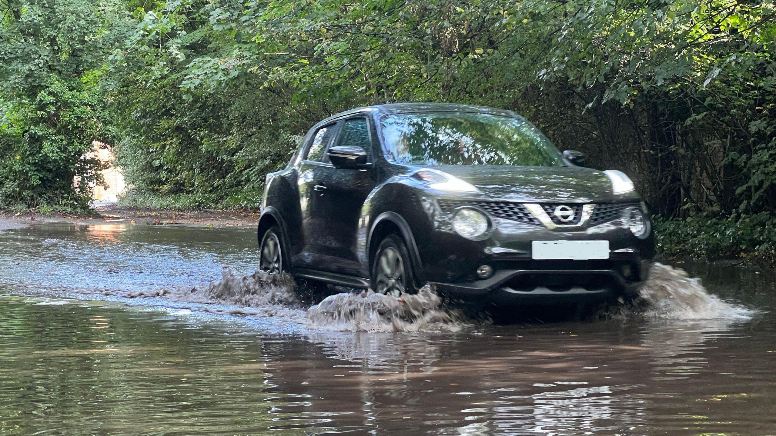 A black car driving through the flood water on the road with trees in the background.