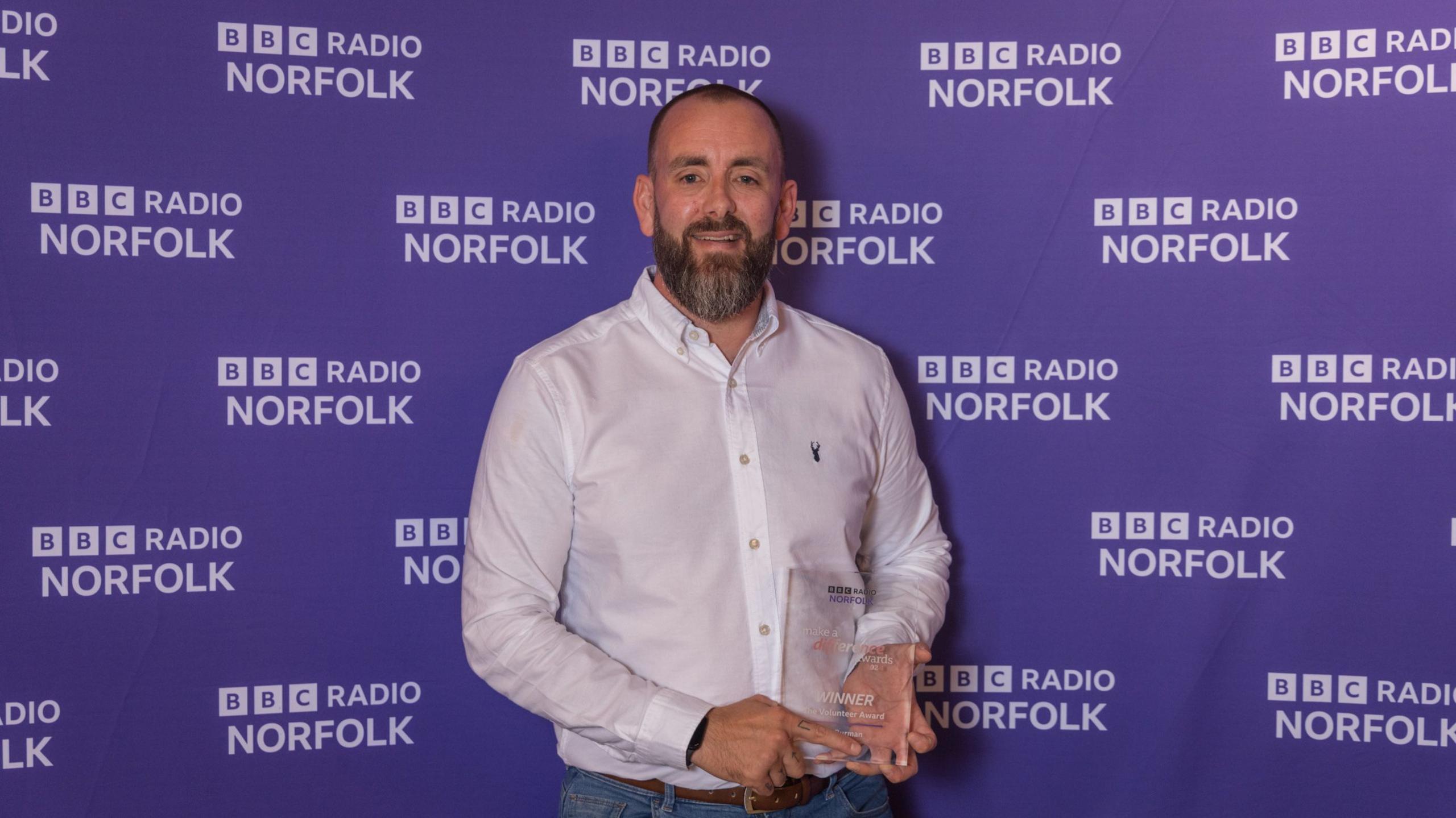 A man with a dark brown beard wearing a white shirt and holding an award while standing in front of a purple BBC Radio Norfolk backdrop