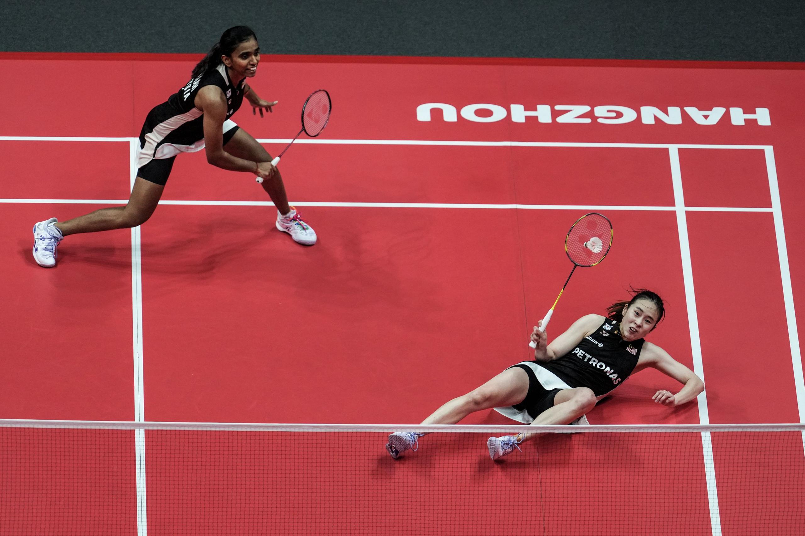 Two badminton players are on a bright red court during a doubles match. One player is standing near the net, holding a racket and appearing ready to return a shot, while the other player is lying on the court with a racket in hand, seemingly after a dive. The court markings and the word “HANGZHOU” are visible on the surface.