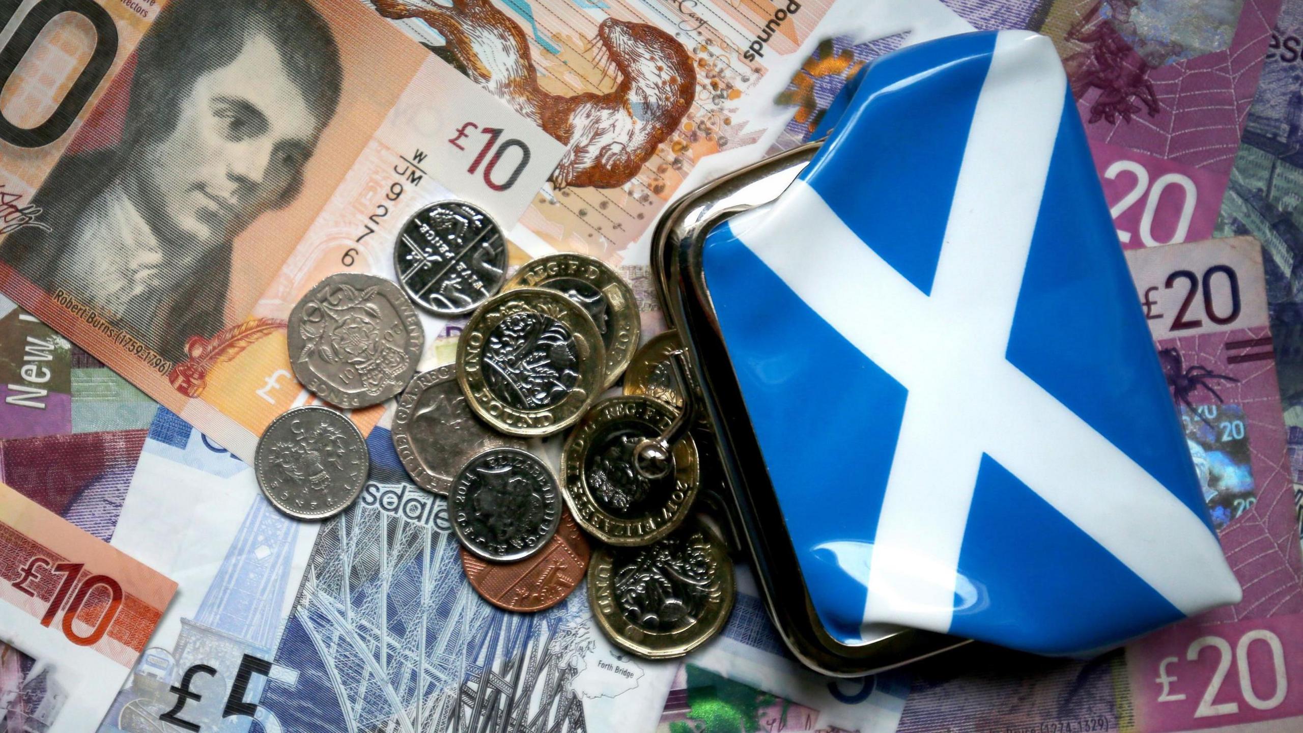 A selection of Scottish money, including 1p, 5p, 20p, £1, £5, £10 and £20 notes spilling from a purse with a saltire on it.