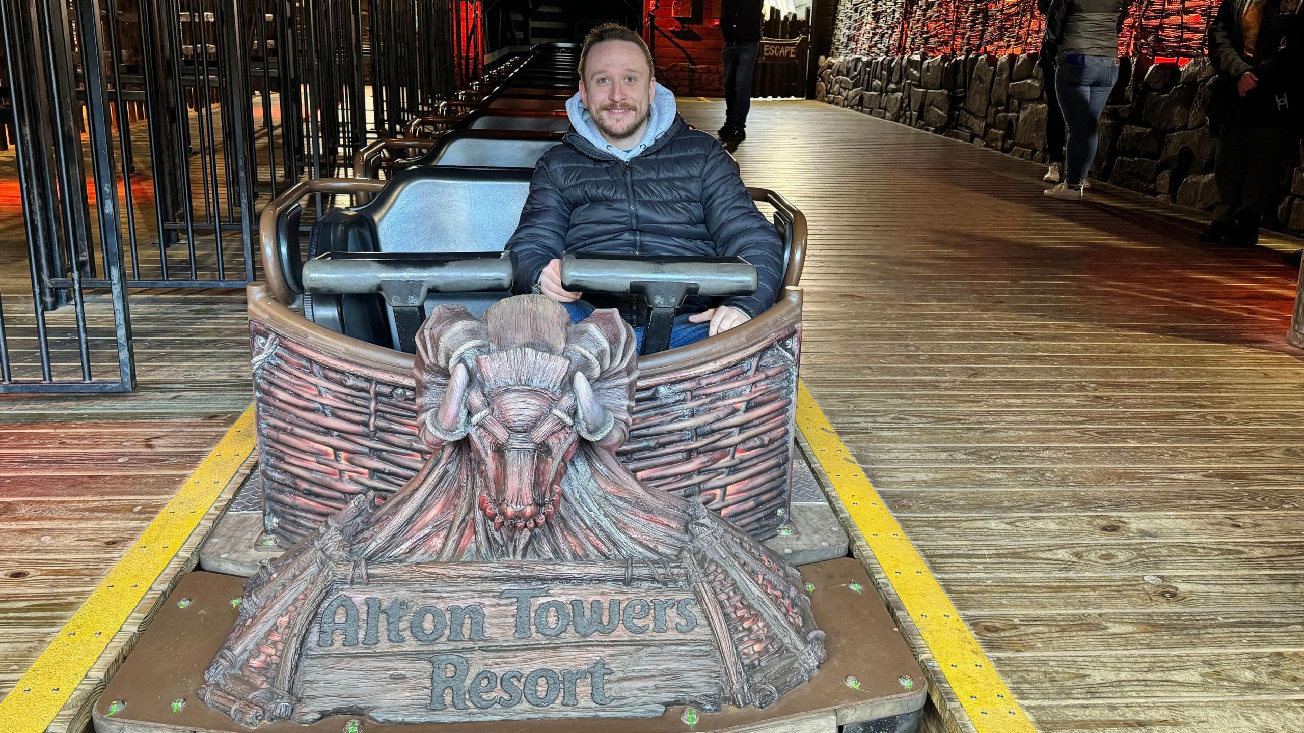 Mr Evans, wearing a black coat, sitting in the cart of a rollercoaster at Alton Towers.