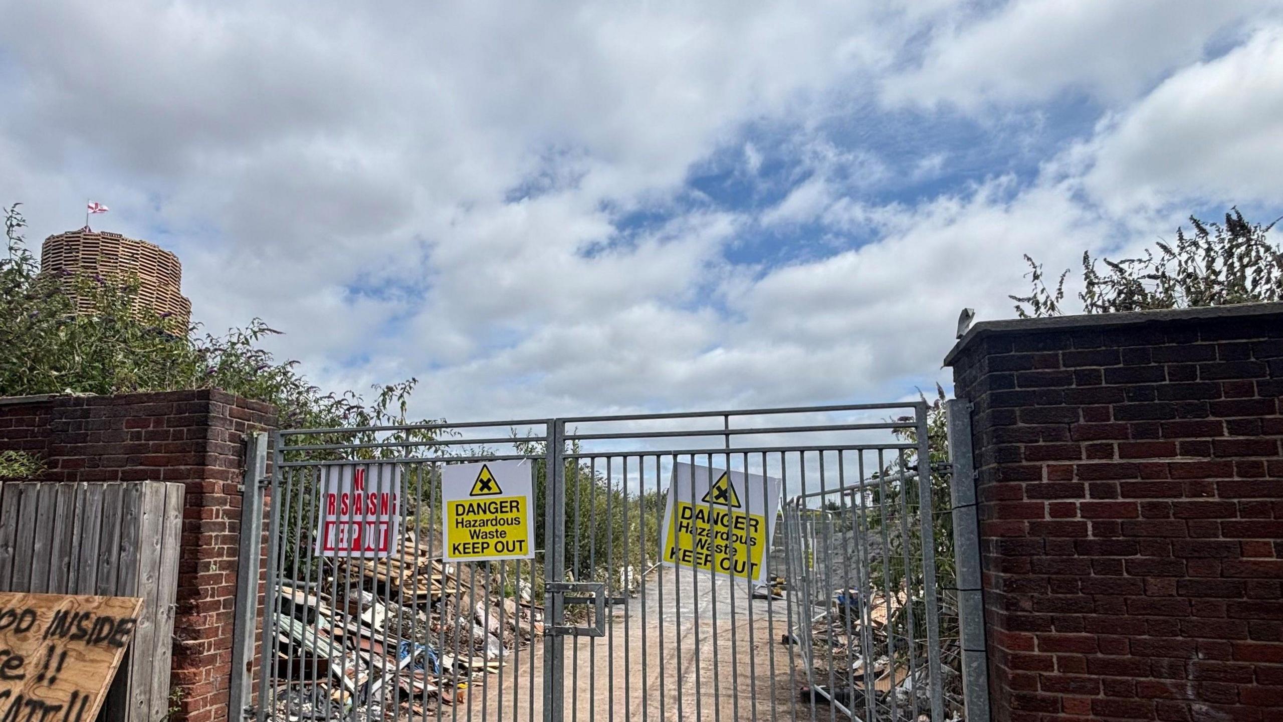 The bonfire site, which has been closed off by metal gates with signs reading "DANGER HAZARDOUS WASTE KEEP OUT", in the corner you can see the bonfire.