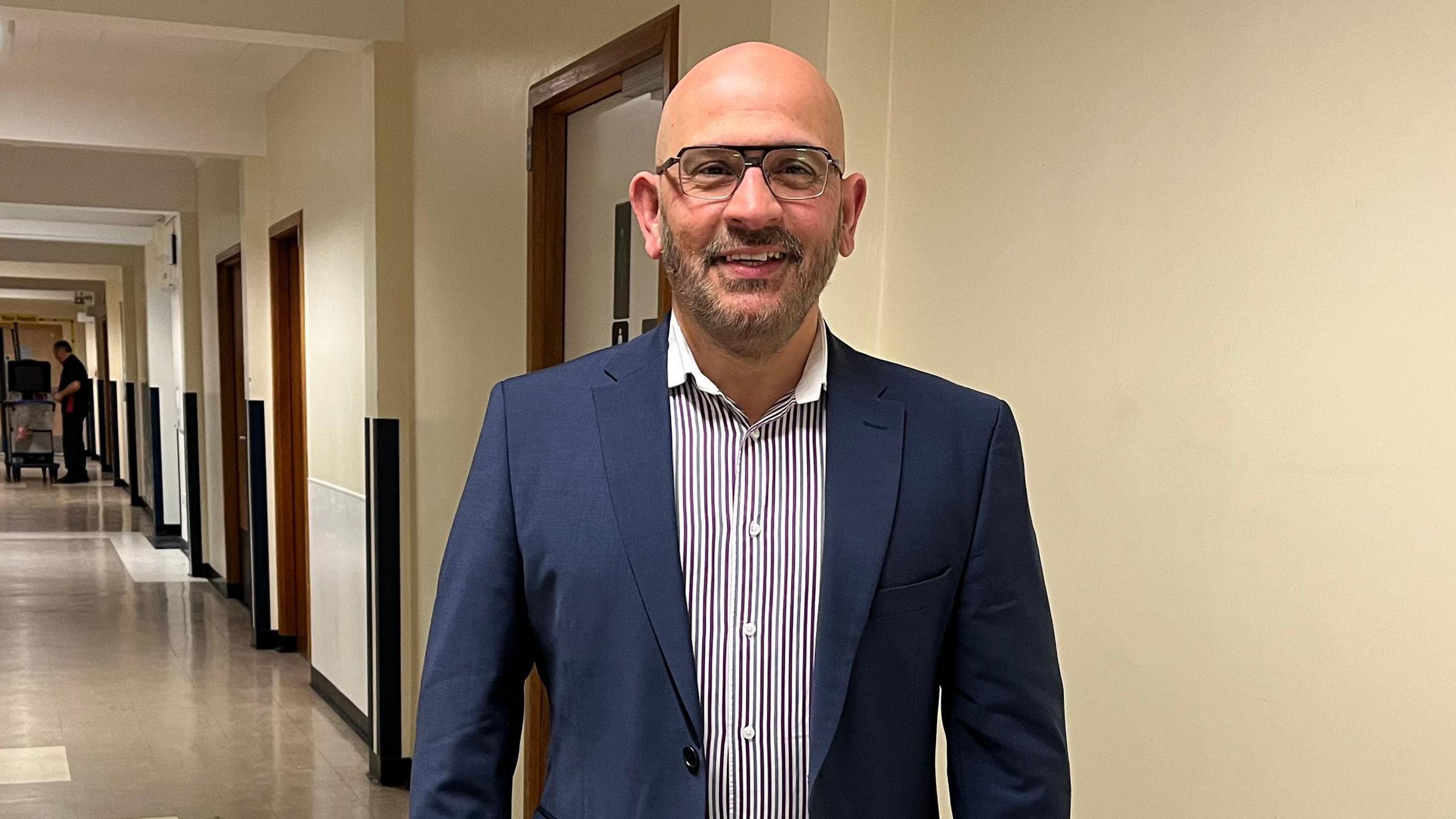 Syed Hyder in a corridor of the hospital, smiling into the camera. He has a bald head, black and grey beard and is wearing glasses. He is wearing a blue suit and striped shirt.