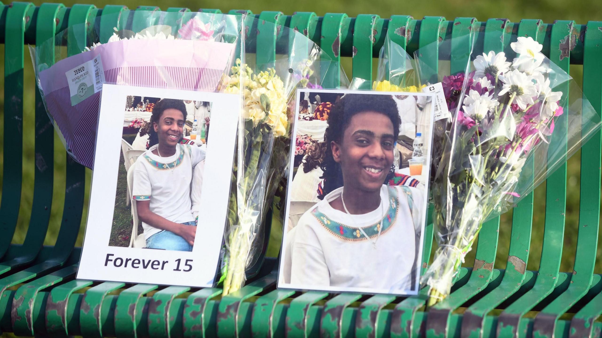 Tributes to Amen connected  a greenish  bench. There are pictures of him smiling and bunches of flowers.