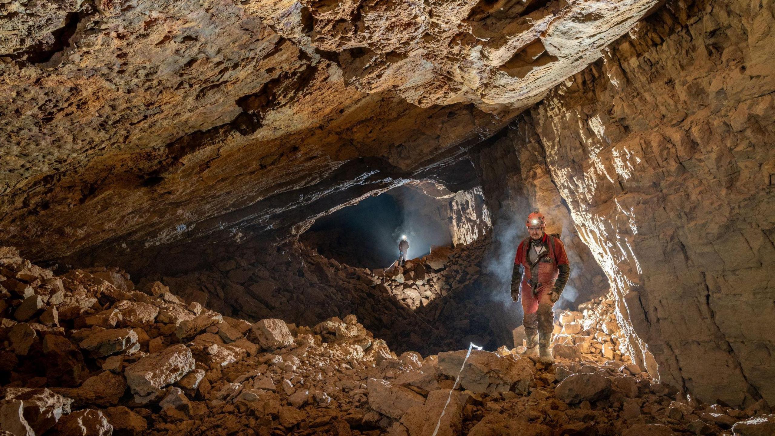 Two cavers are seen, one behind the other, exploring a large cavern underneath English Bicknor in the Forest of Dean. Around them are large slabs of reddish rock and their headlamps are illuminating the space