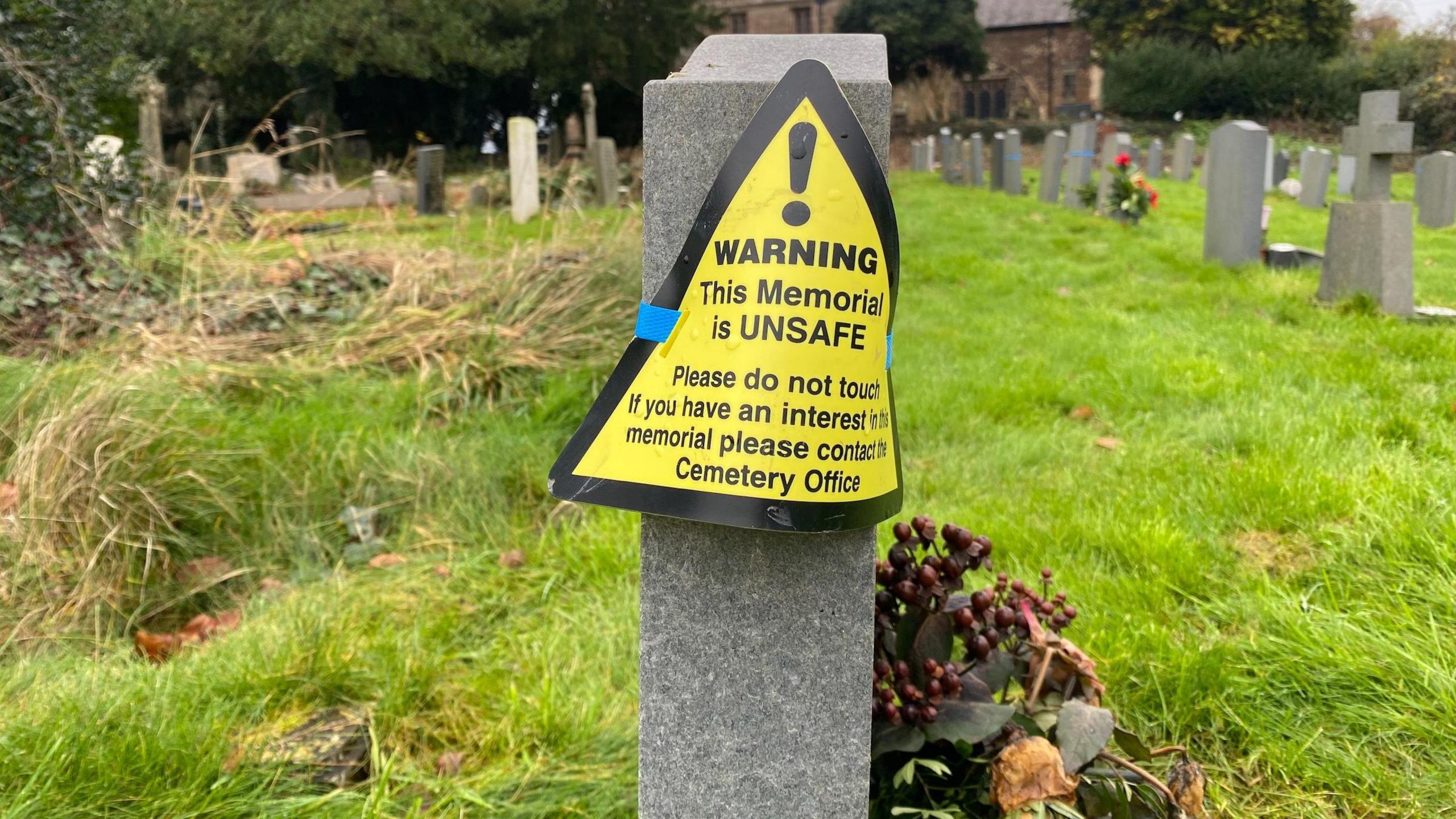 The side of a grave that has a yellow sign strapped to it, marking it as unsafe. There are a number of graves in the background.