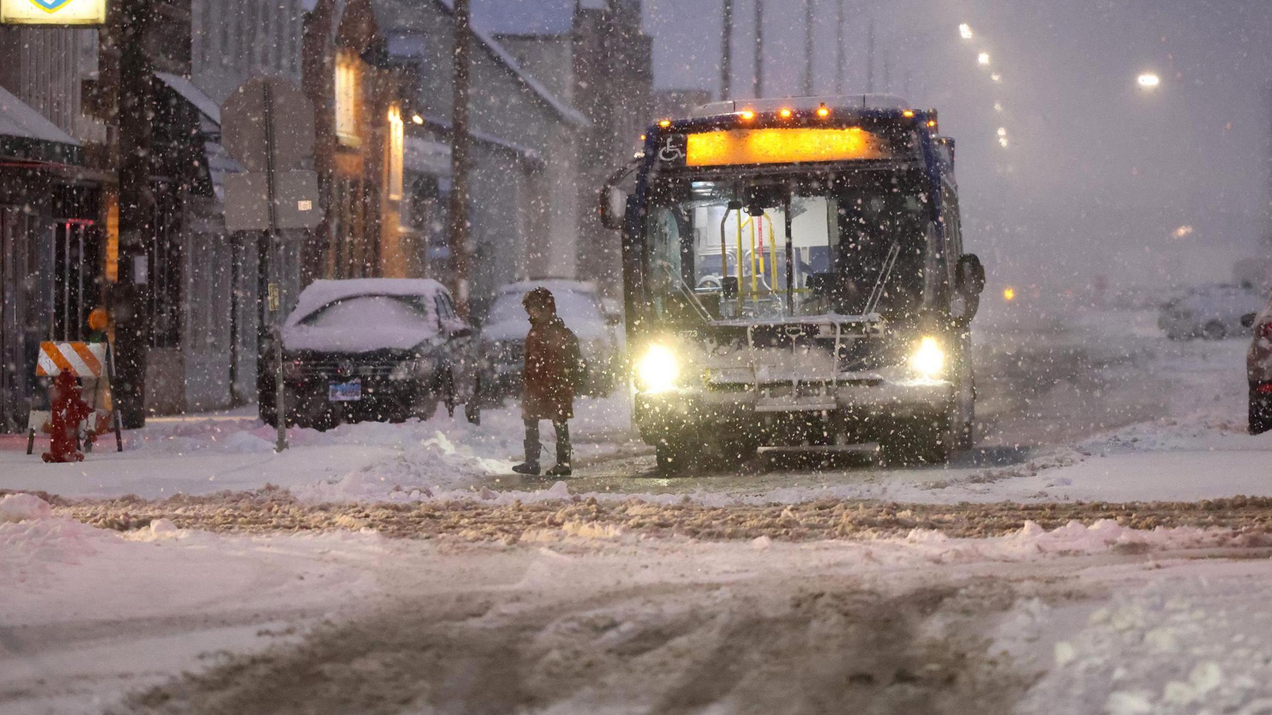 A bus drops off a commuter in Chicago, Illinois during a winter storm. Snow is visible falling and piled up on the road.