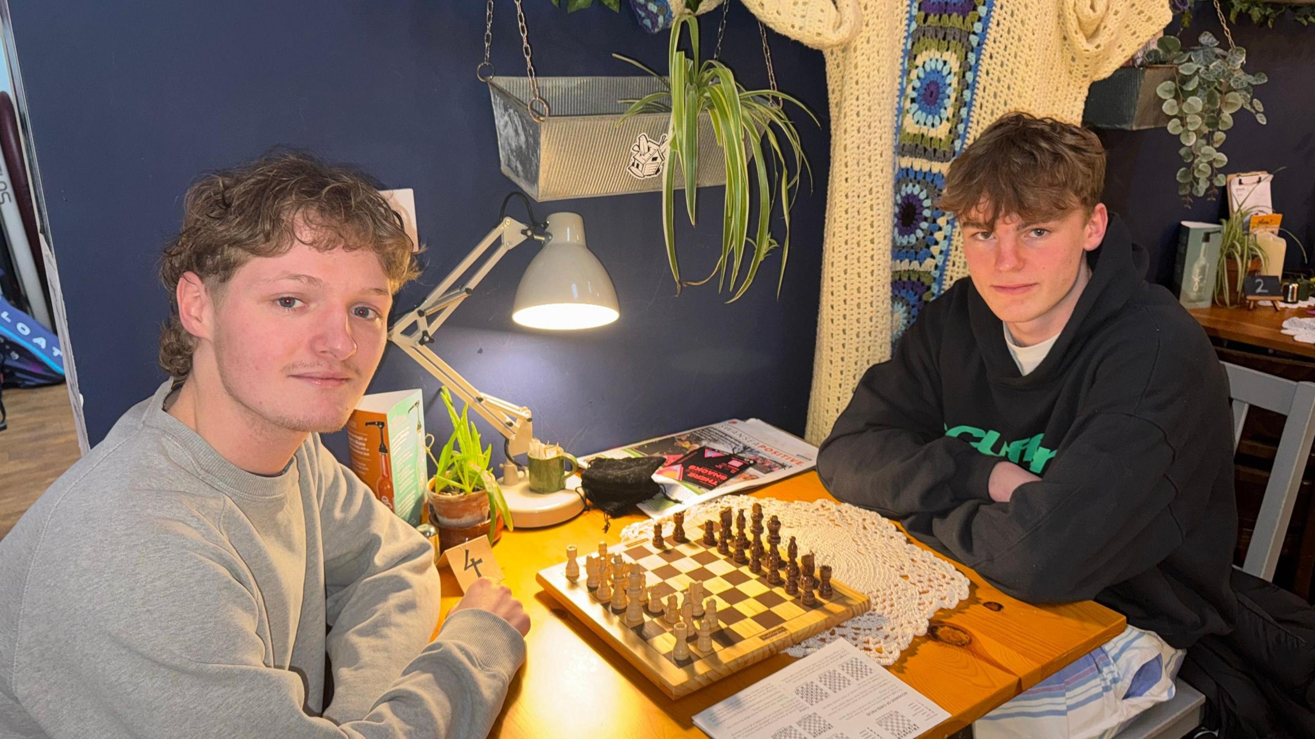 Will and George sat at a table, with a chess board in front of them. One of the students has a faint moustache and curly hair, and wears a grey sweater, and the other has short darker hair, and is wearing a black hoodie. There is a navy blue wall behind them with trailing plants in two hangers, and a crochet wall hanging in the background. 