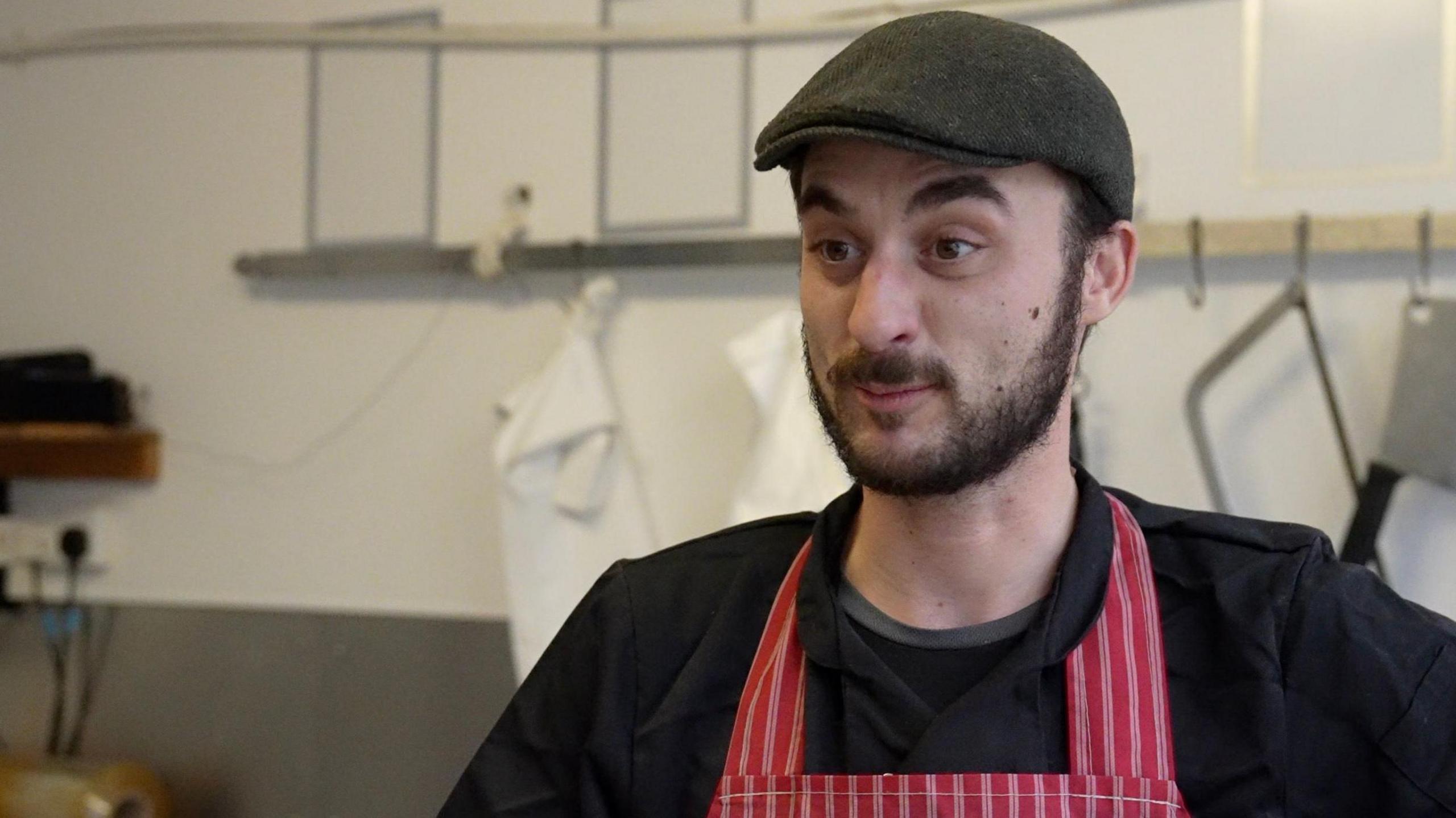 A butcher stands in his shop, which has a white a grey wall with hooks for white aprons. He has a dark brown beard and is wearing black overalls, a black cap and a red-and-white striped butcher's apron. 