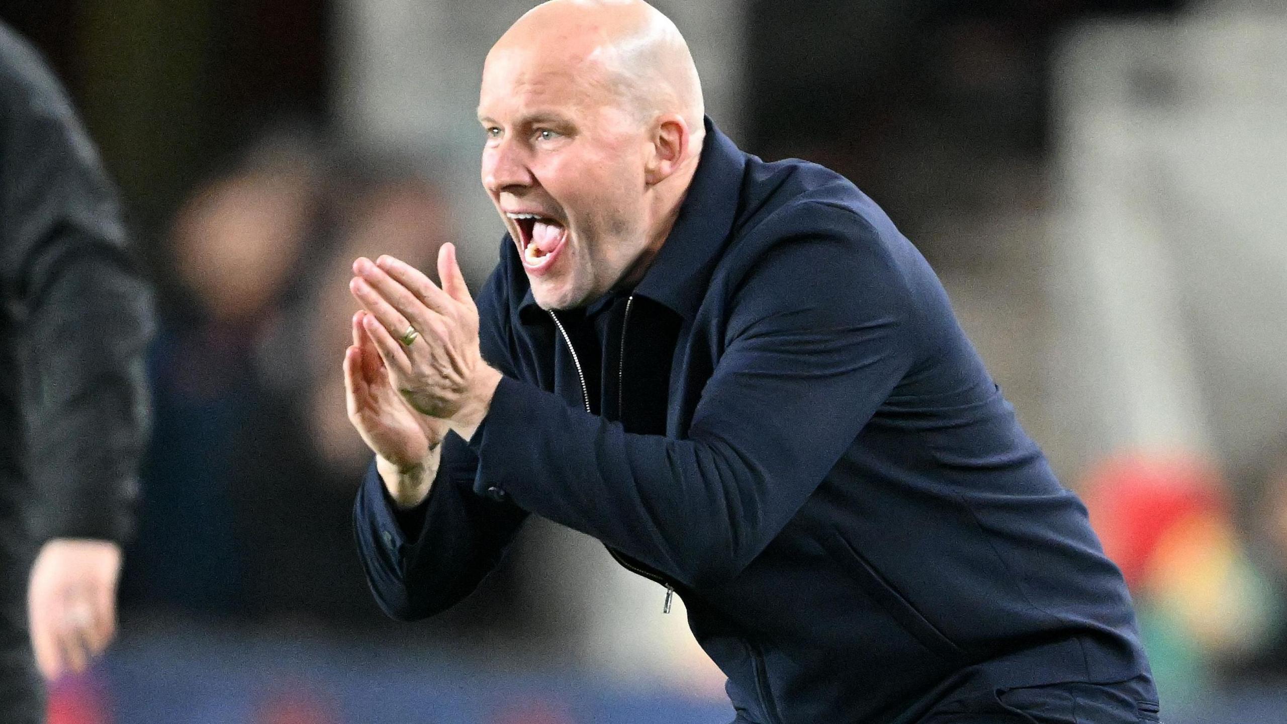 Henrik Pedersen crouches on touchline, hands clapping encouragement as he shouts instructions, wearing all navy blue trousers and zipped warm jacket.