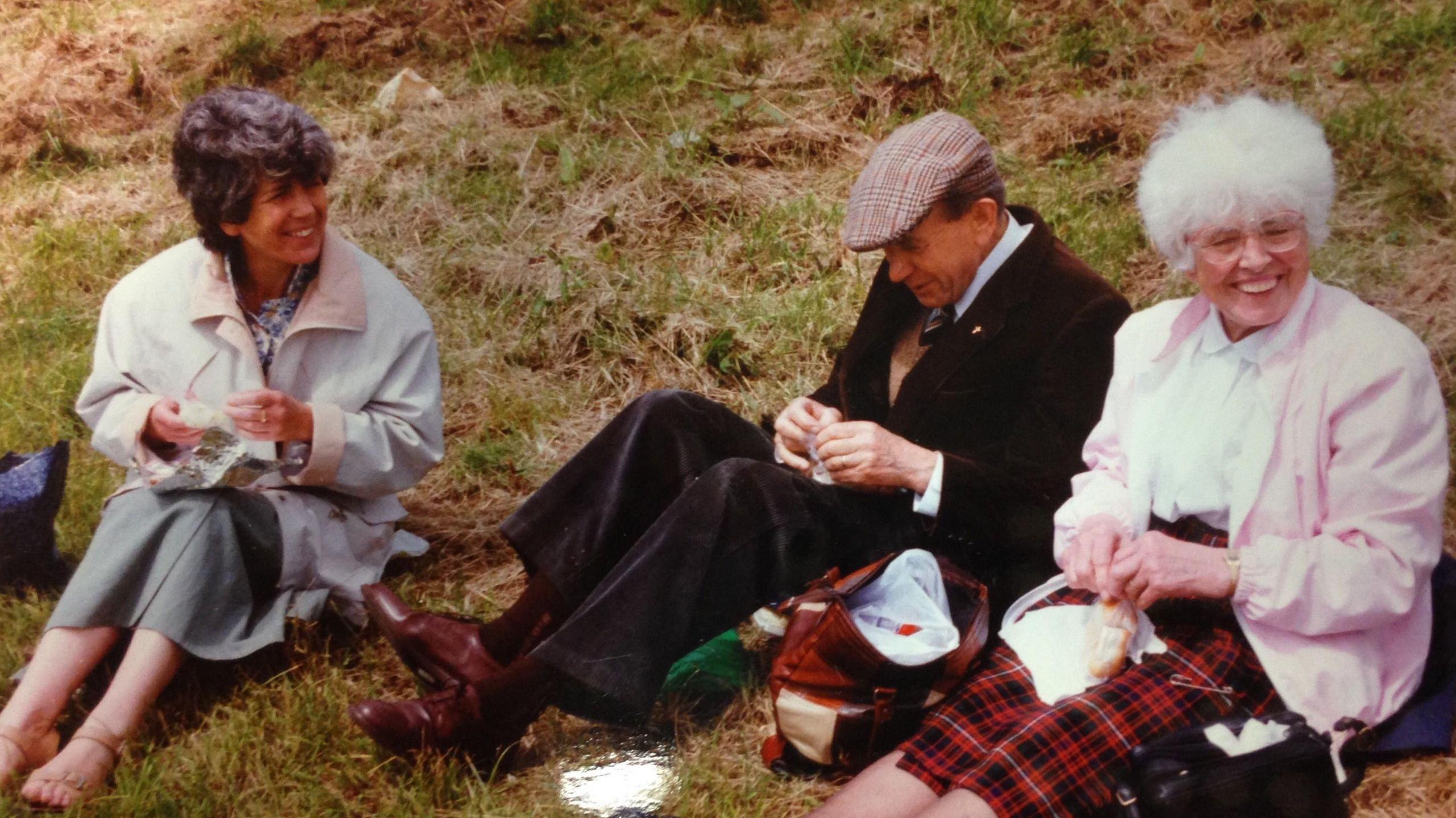 A woman with dark curly hair sitting on grass along with an elderly man in a flat cap and an elderly woman with white hair and a pink jacket. They are having a pic nic.