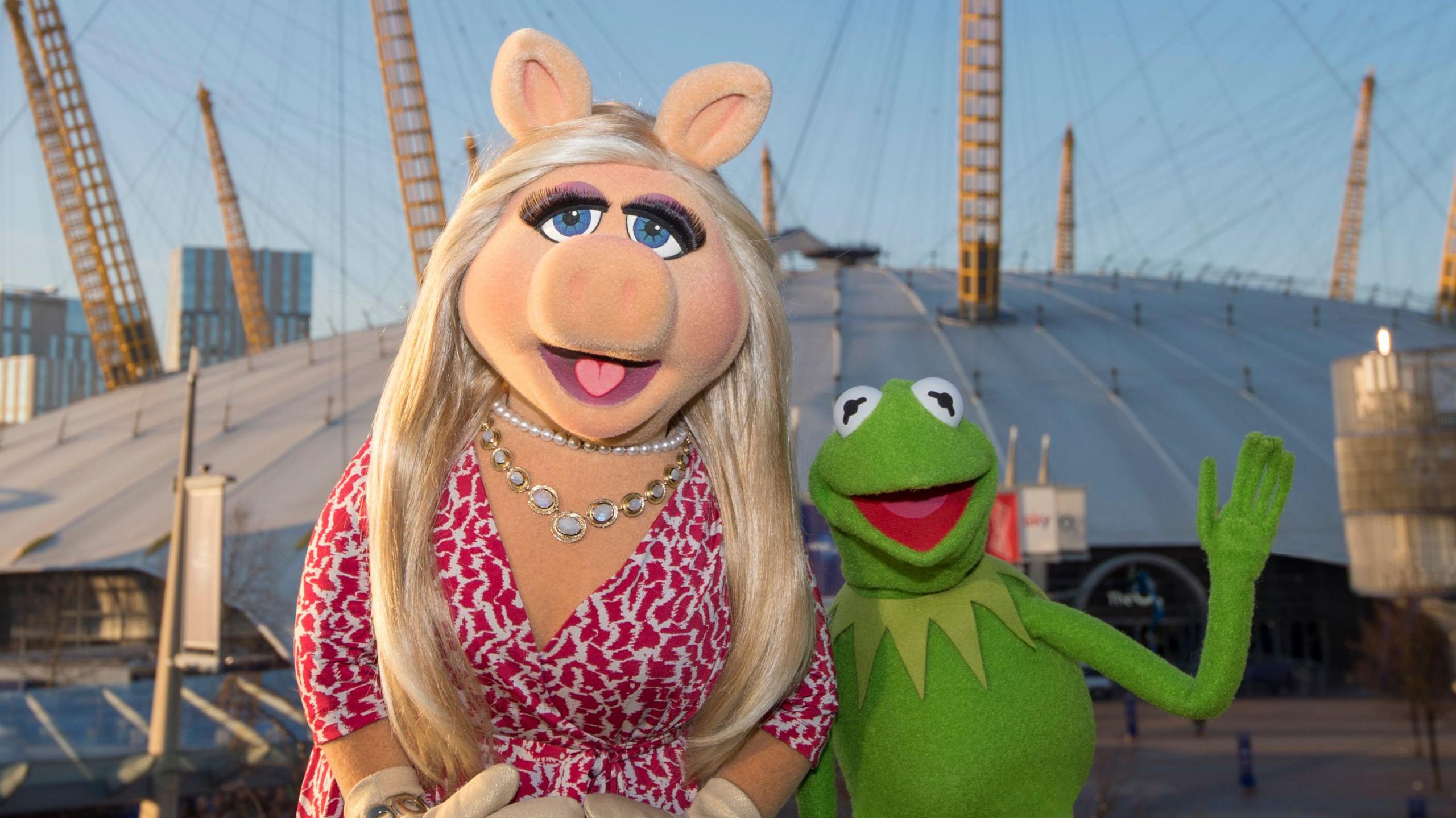 Miss Piggy wears a white and pink dress and silver jewellery. She stands beside Kermit the Frog who is waving. The pair are standing in front of The O2 in London.