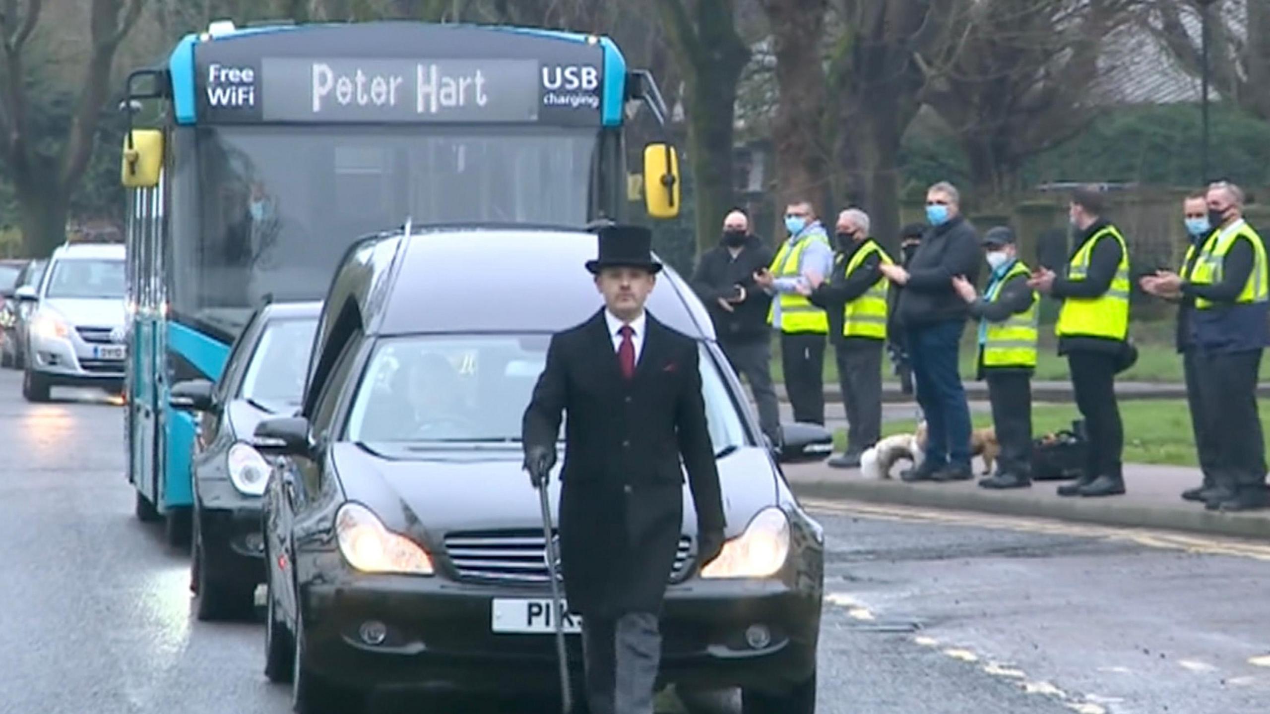 A funeral director leads a cortege of vehicles including a bus with the name Peter Hart on its screen while bus drivers line the street in hi-vis jackets applauding the cortege