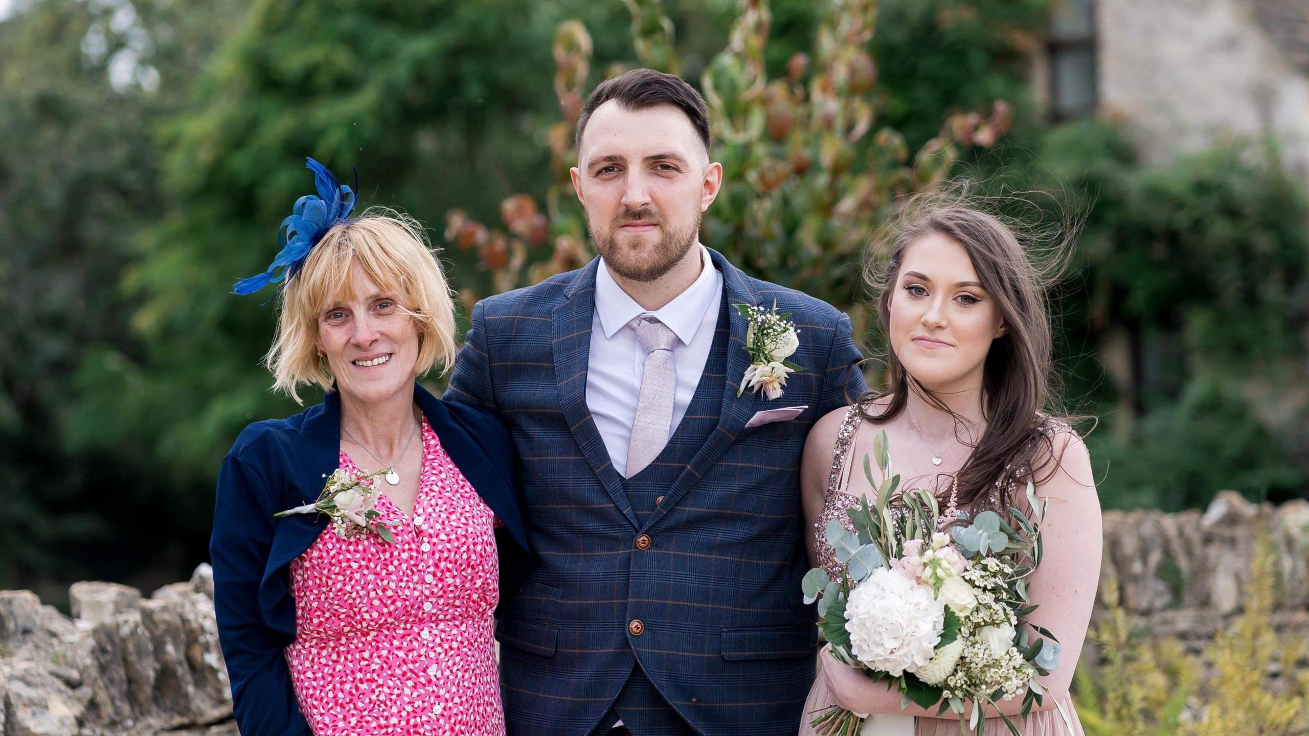 Becky stands with her brother and mother on her right. They are all in smart wedding attire. Her mum is in a pink dress with small white flowers and a navy light jacket. There is a navy fascinator in her blonde hair. Becky's brother is in the middle, wearing a navy three-piece suit, an off-white shirt and a cream tie. He has brown hair and a slightly longer than stubble beard.
Becky is in a light pink sparkly dress and is holding a big bouquet of flowers.