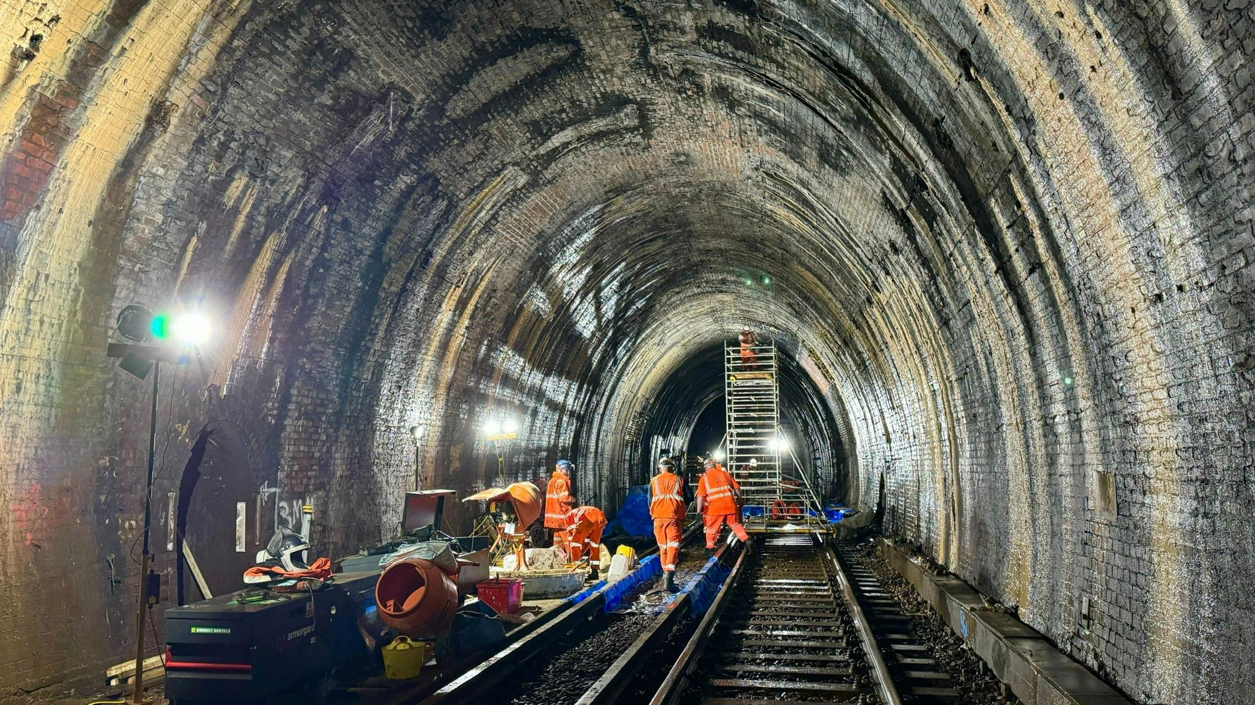 The Blackheath tunnel inspected by four men in orange jackets plus another using ladder. Equipment lays on the tracks to the left.
