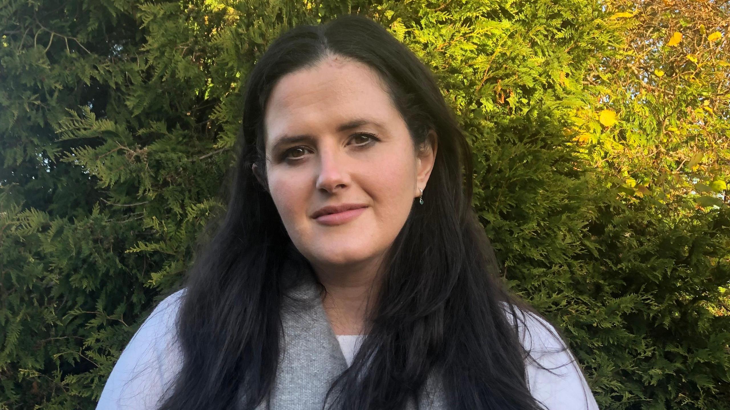 Claire Sugden, a woman with long, straight black hair, looks to the camera while standing outdoors on a sunny day.   She is wearing a white long-sleeved top and a grey scarf.  There are evergreen trees in the background. 