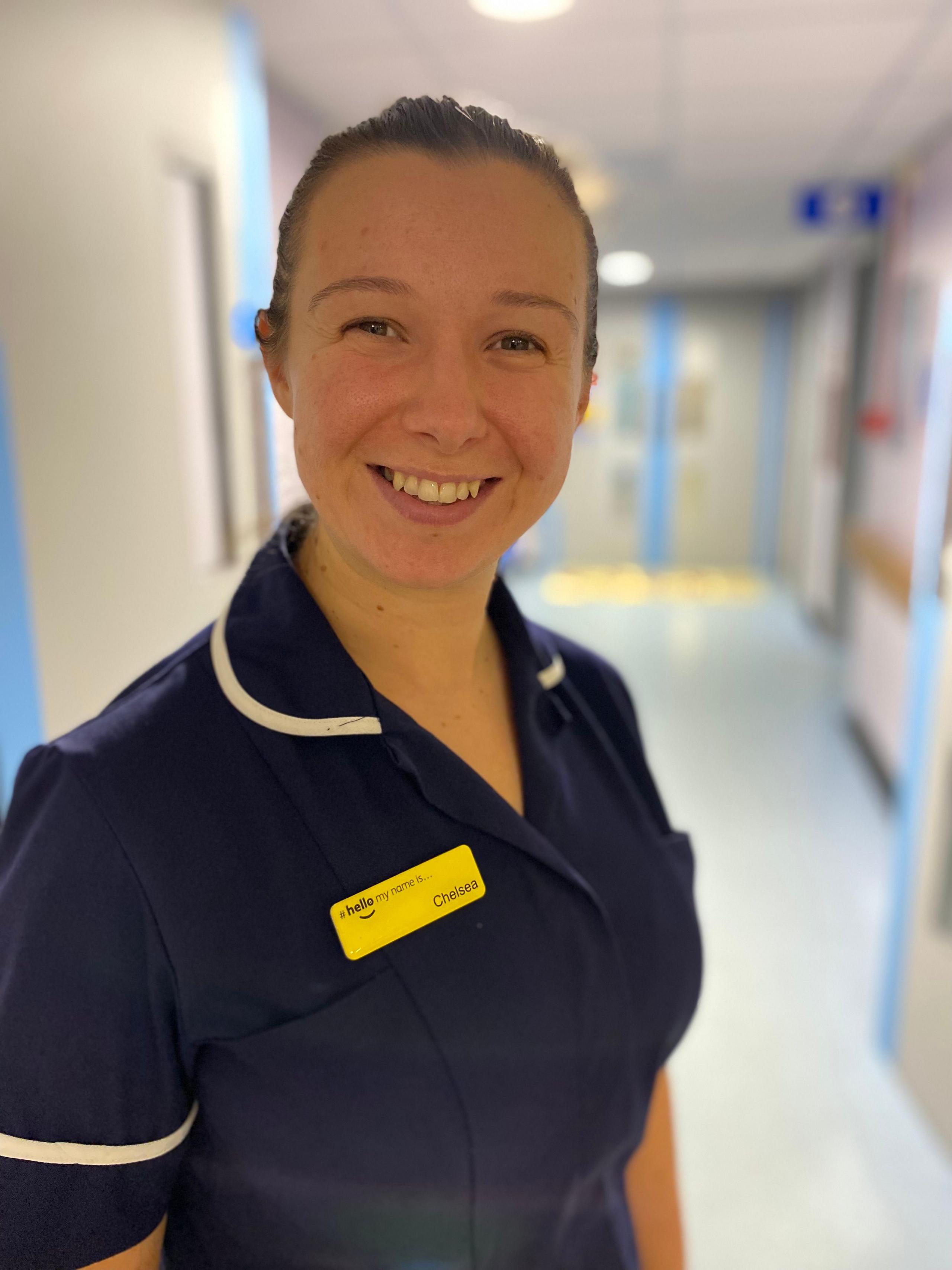 Chelsea Spencer. She is dressed in her blue nurses' uniform, with yellow name tag, and is standing in a hospital corridor. 