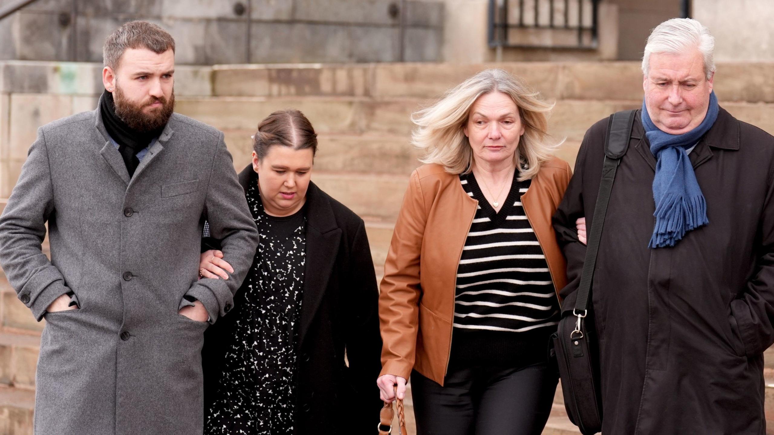 A group leaving the court. From left to right: A tall man in his 30s with a brown beard, wearing a long straight grey coat. A woman of a similar age, up to his shoulders, wearing a black dress with some white patterns and a long black coat. A blonde woman in her 50s, wearing a black and white striped top and brown leather jacker. A man who looks in his 60s with white hair, wearing a long black coat and blue scarf with a black satchel over his shoulders. Each couple has their arms linked.