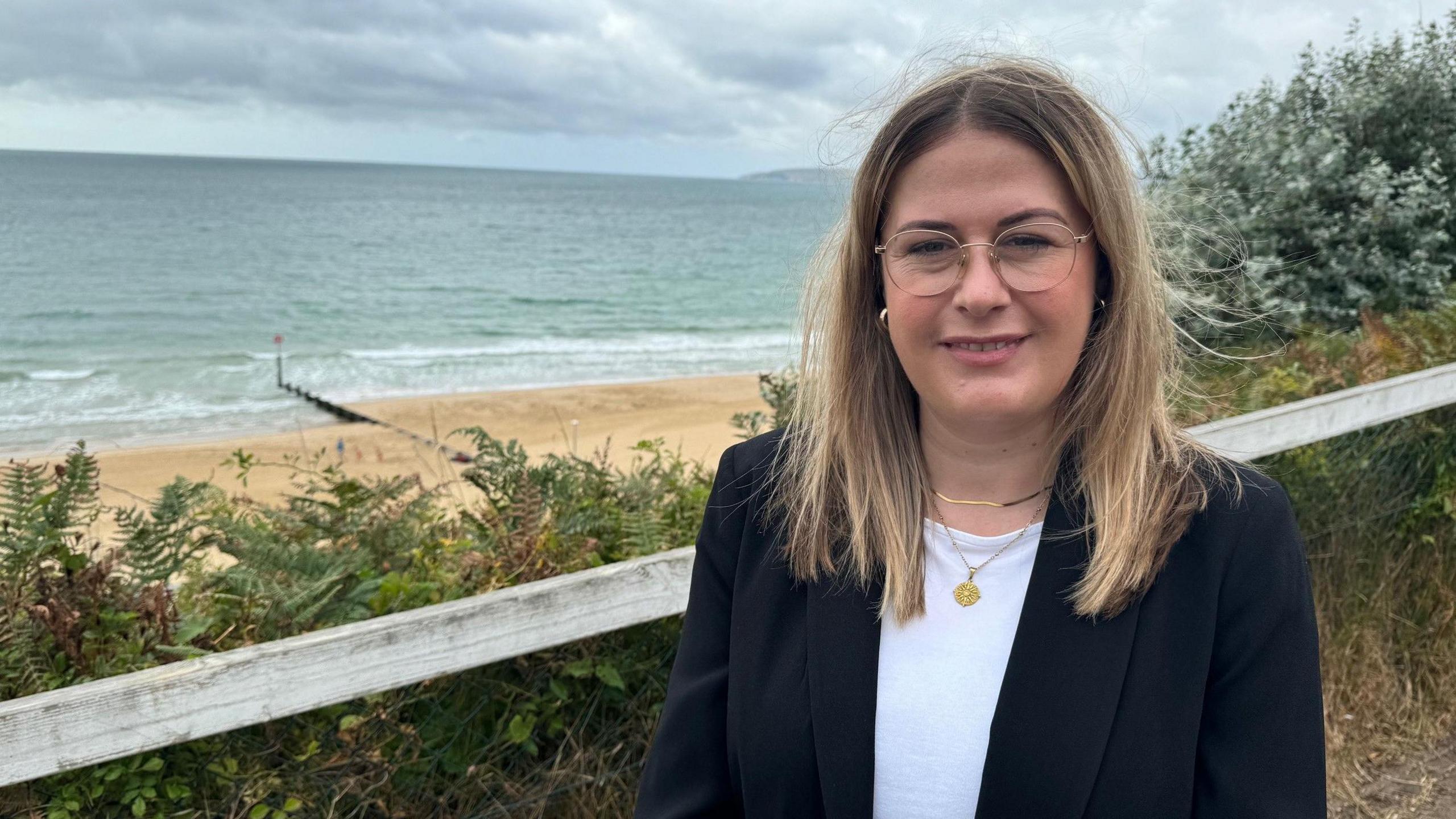 Millie Earl standing on the seafront. She has long fair hair and round glasses and is wearing a white top and black blazer. In the background is the beach and the sea