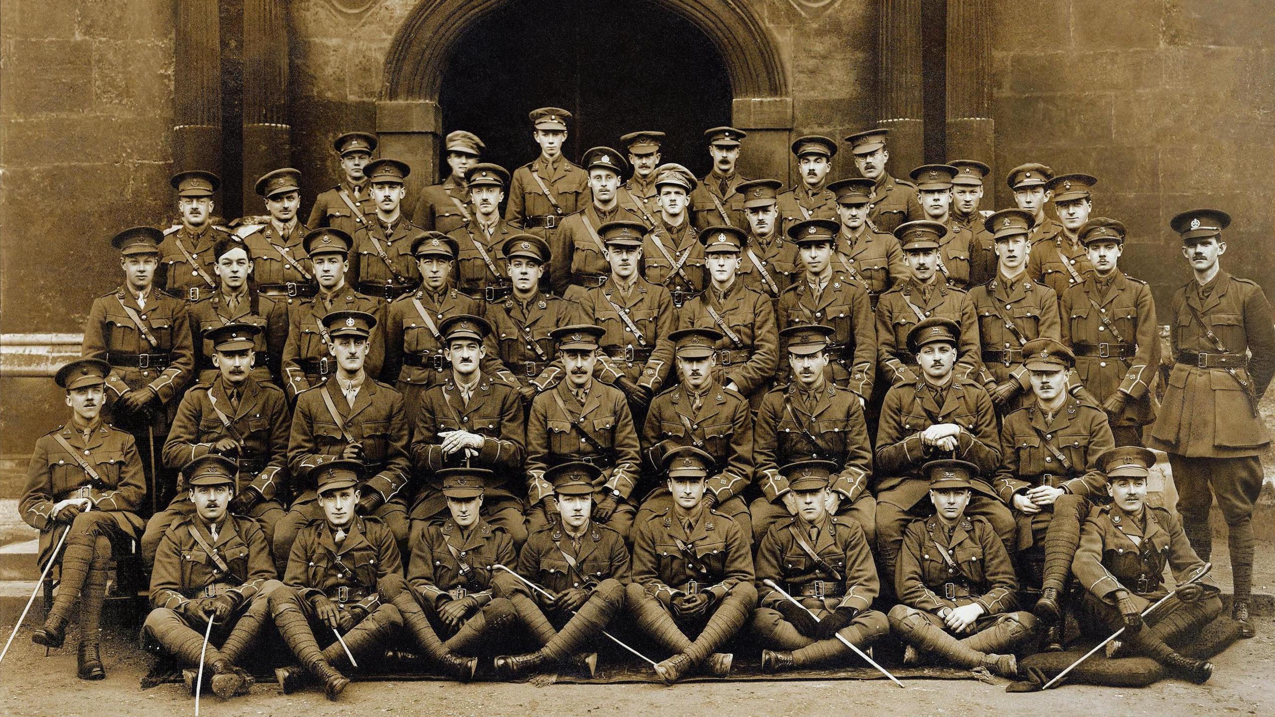 Sepia-toned photograph of 48 men in military uniform. They are standing in fofront of an archway in military outfits. The men are arranged into four rows of increasing height, with the front row sitting cross-legged on the ground.