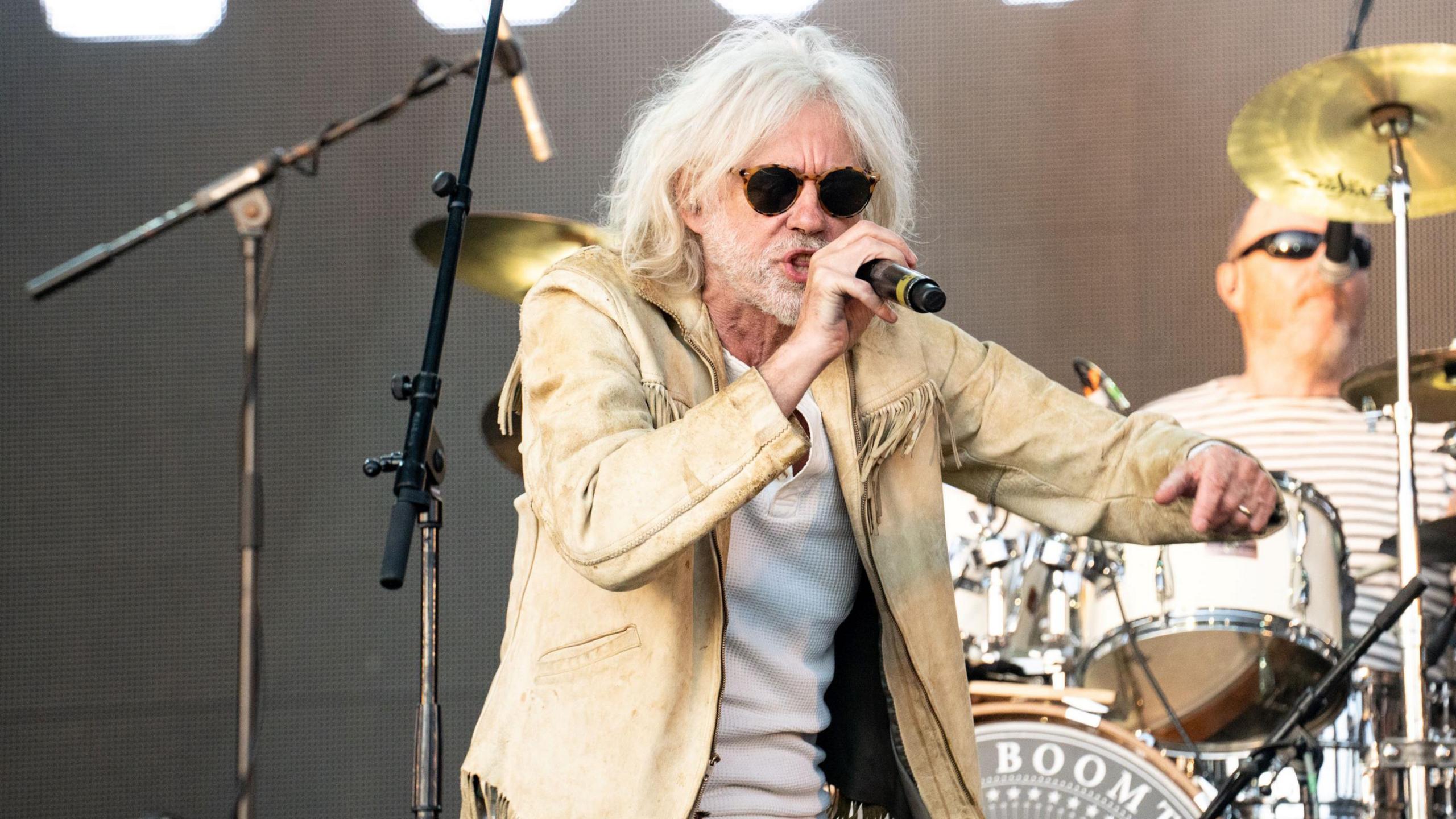 Bob Geldof of The Boomtown Rats singing with his band onstage, wearing a beige suede jacket with tassels