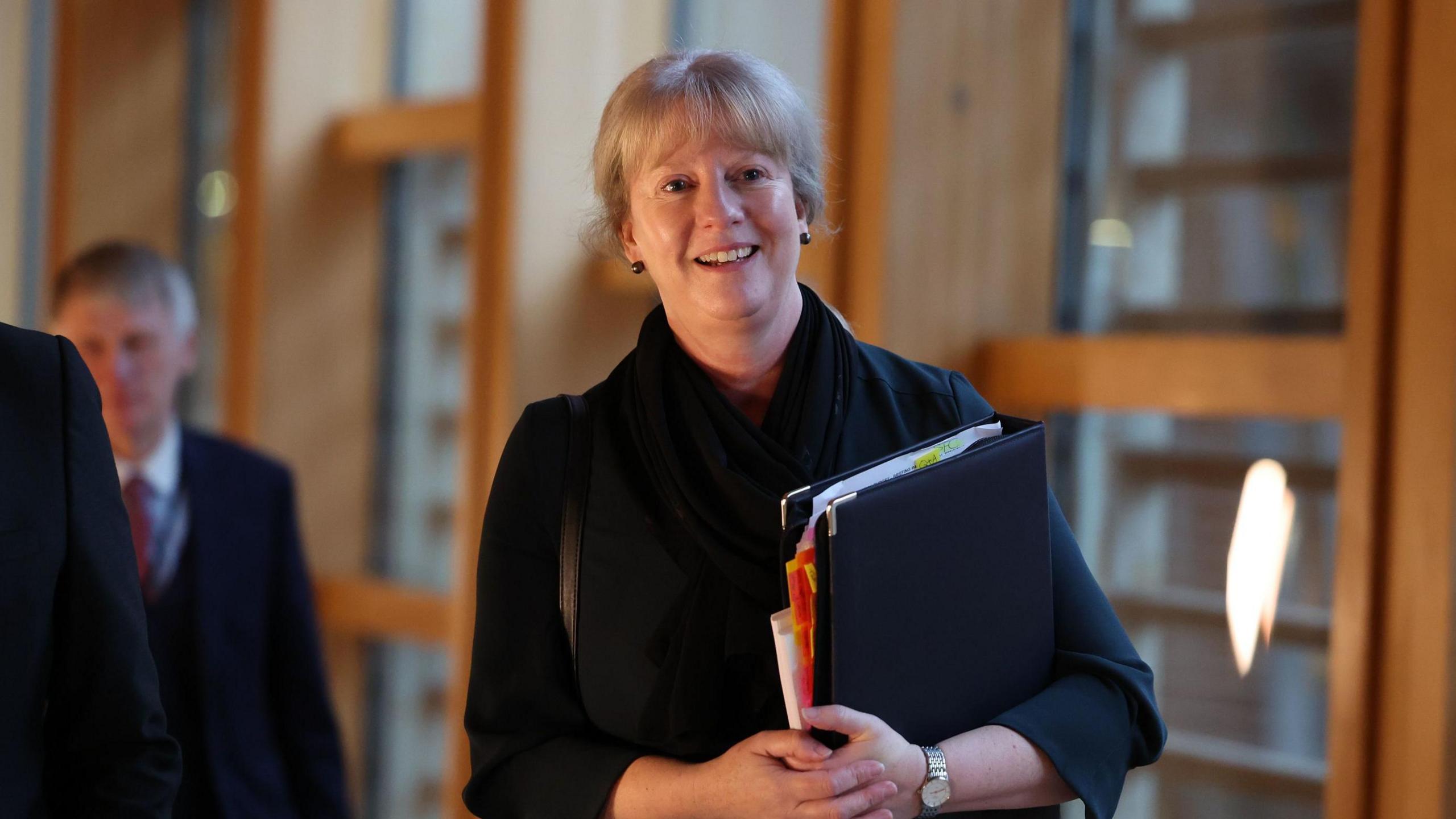 Shona Robison carrying a black folder as she heads into the Scottish Parliament to deliver the budget. She is smiling and wearing a black outfit. 