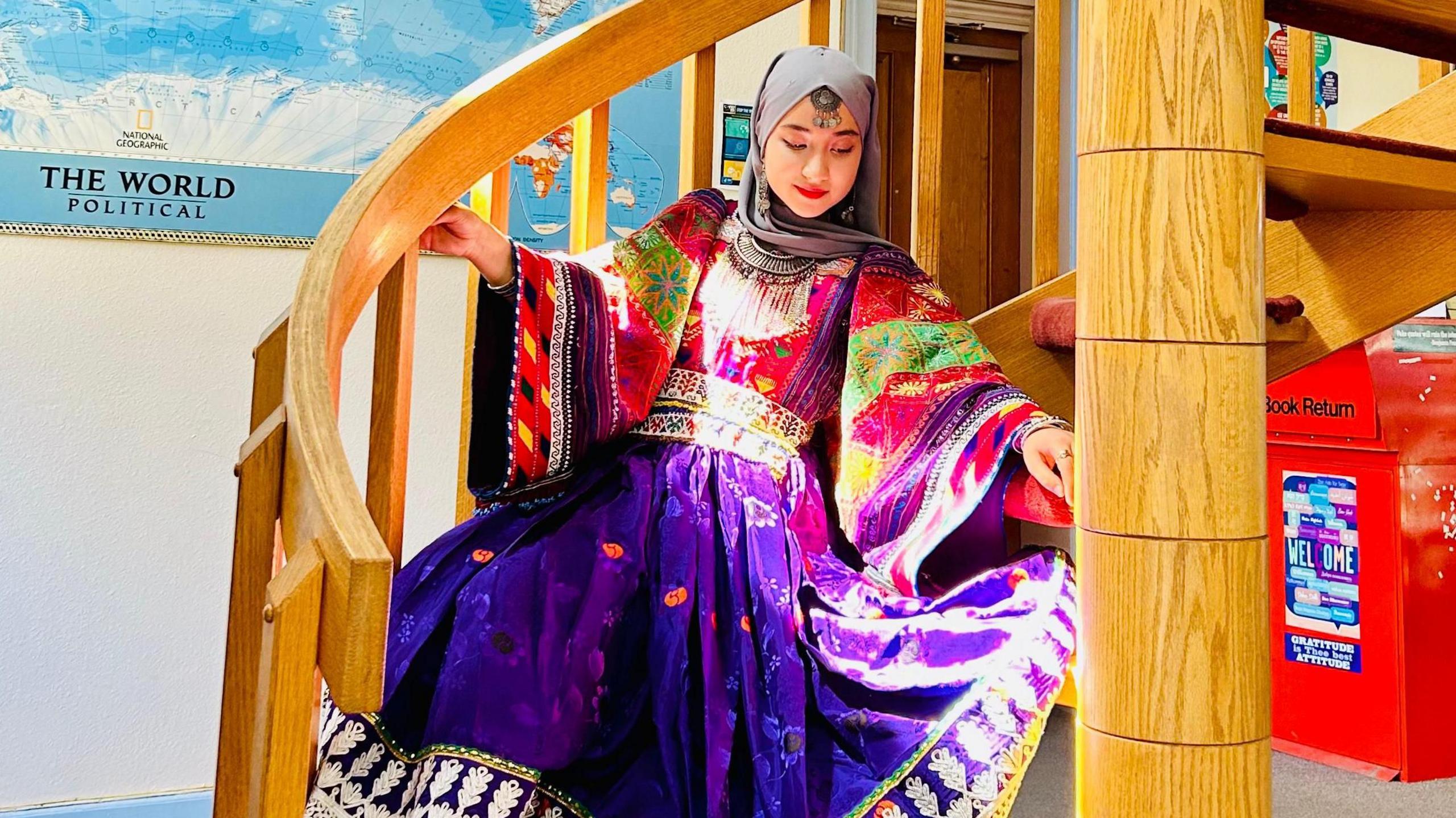 17-year-old Aqdas in traditional Afghanistan outfit, coluored purple, with a world map behind her. Sitting on a brown wooden spiral staircase.