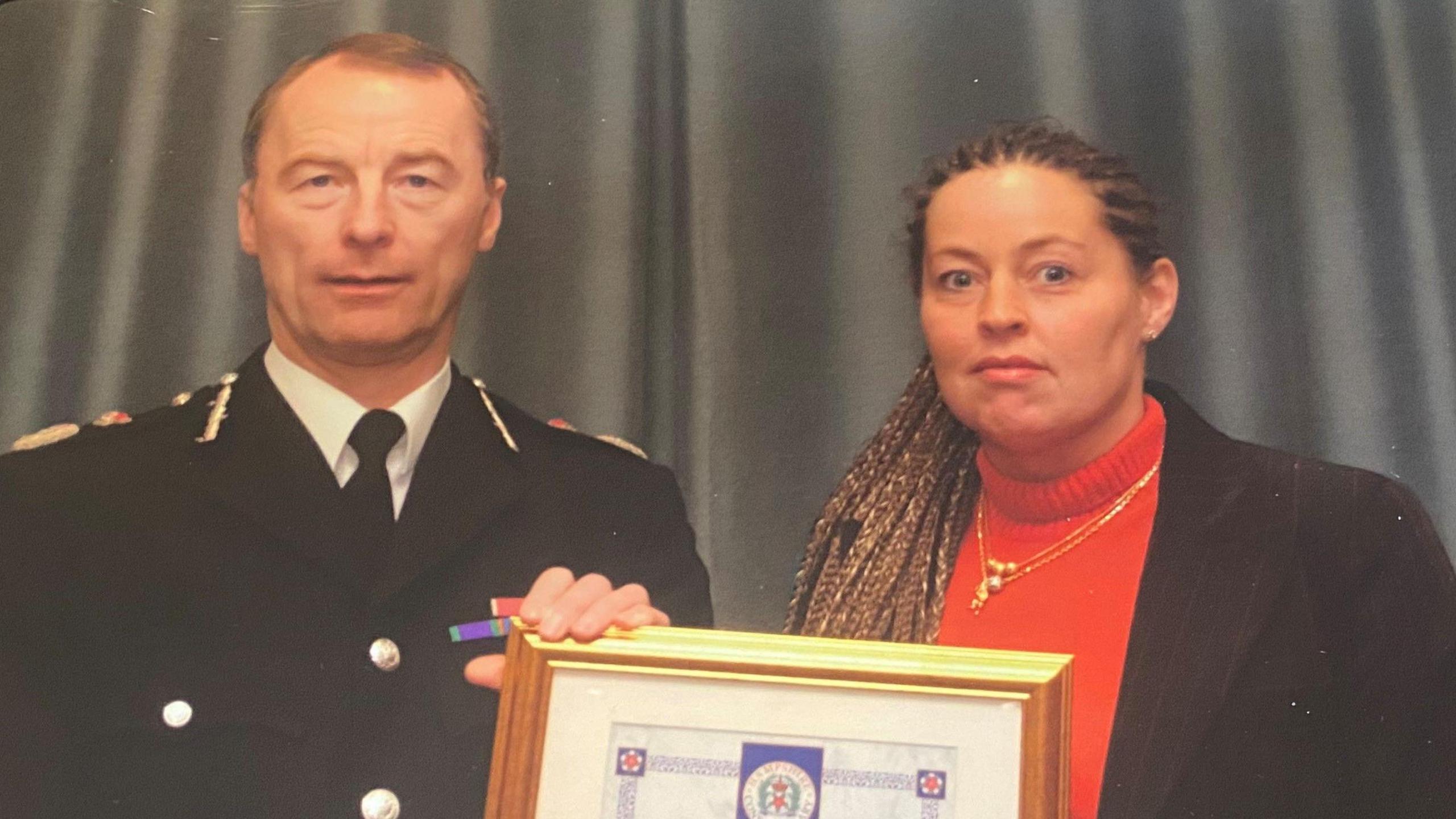 Whendie Backwell, a woman with brown hair in cornrows and plaited into long braids holds a certificate in a gold frame. She is wearing a red jumper. Next to her is an older man with thinning grey hair, possibly high up in the police force. He is wearing a suit.