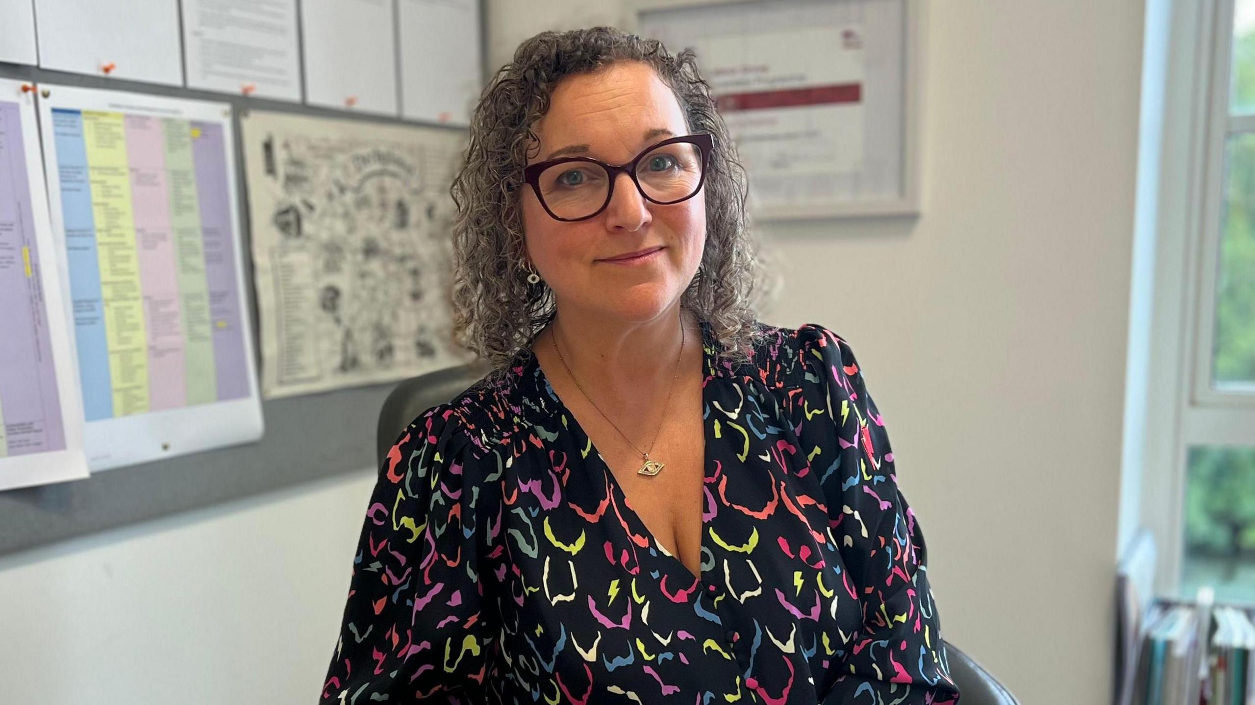 Woman wearing a dark blouse with a colourful abstract pattern featuring shades of pink, green, yellow, and blue and a gold necklace