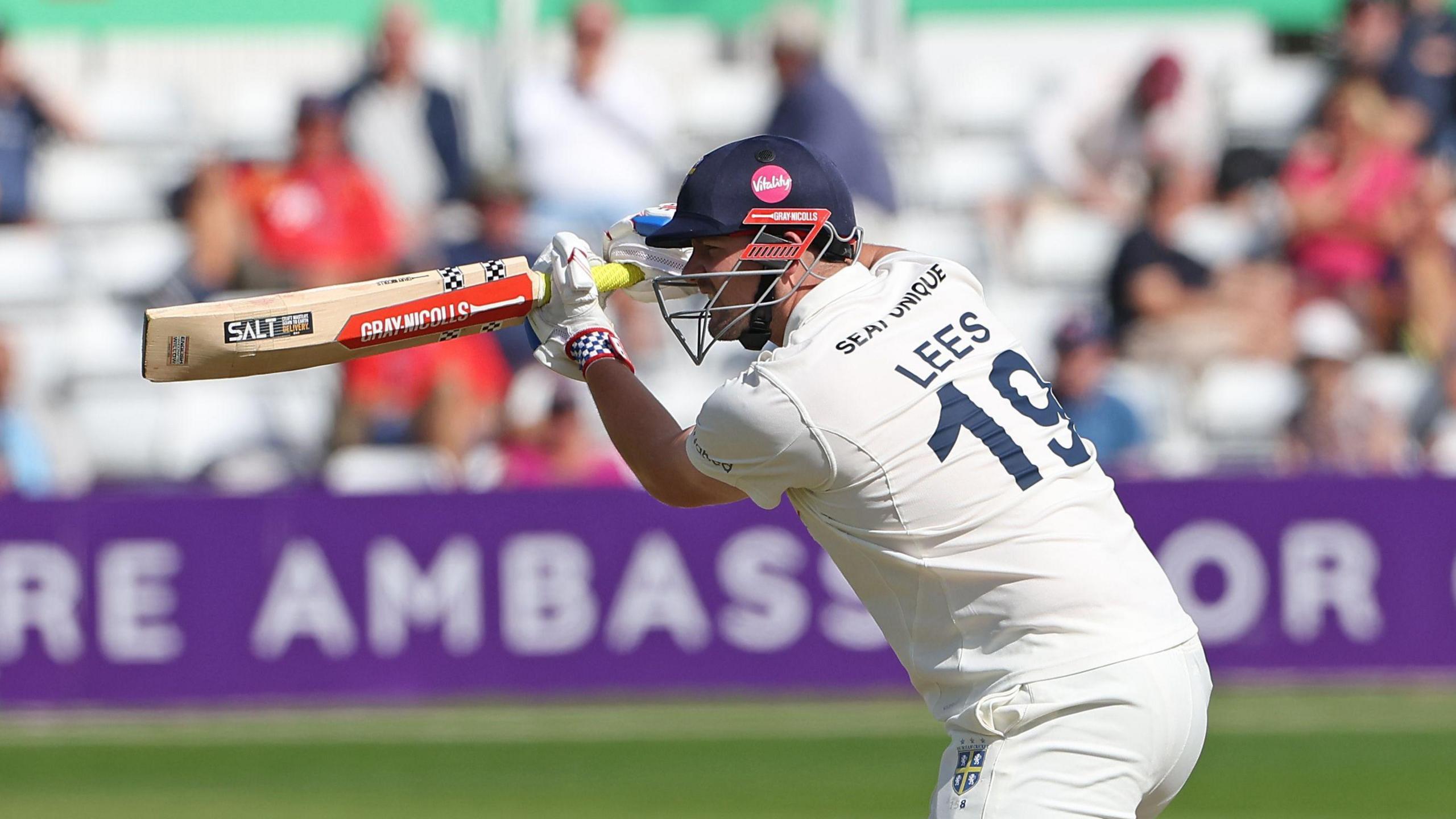 Left-handed batter Alex Lees, wearing "LEES 19" on the back of his white shirt, playing a shot through the offside for Durham, with a helmet on and a high bat.