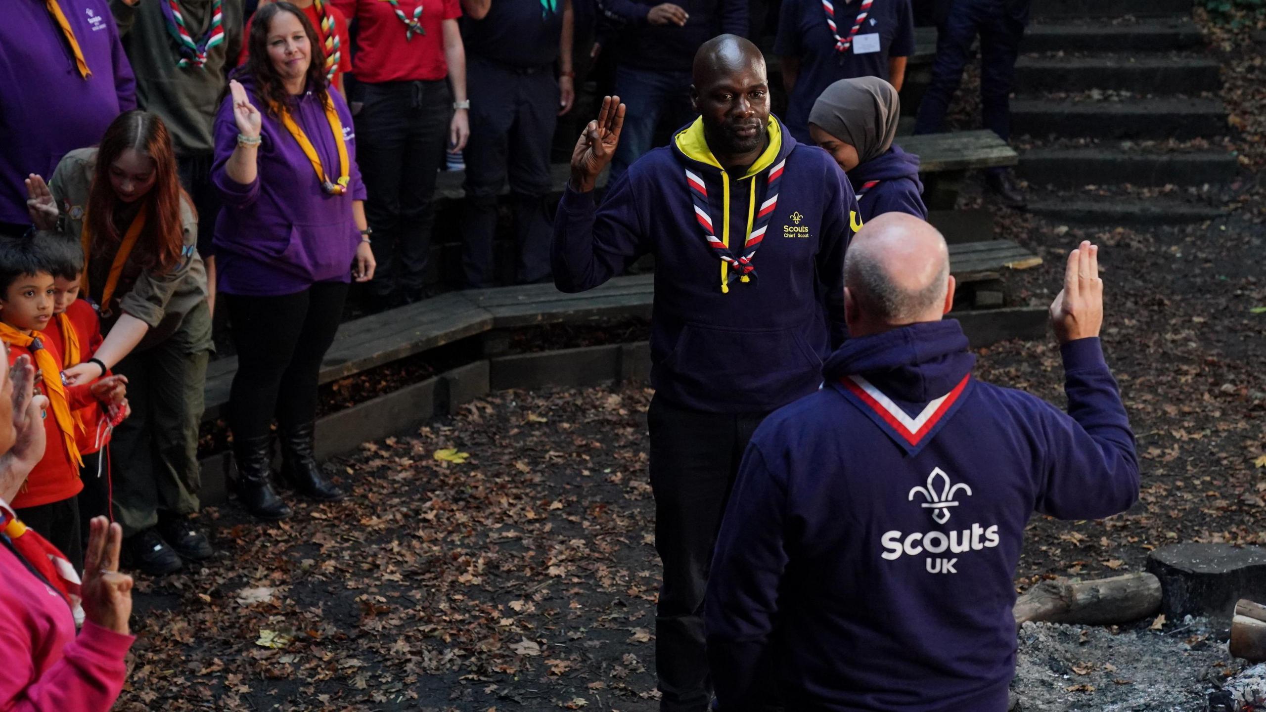 Dwayne Fields giving a Scouts salute in front of a Scout group leader by a campfire while a crowd of people watch on from the side.