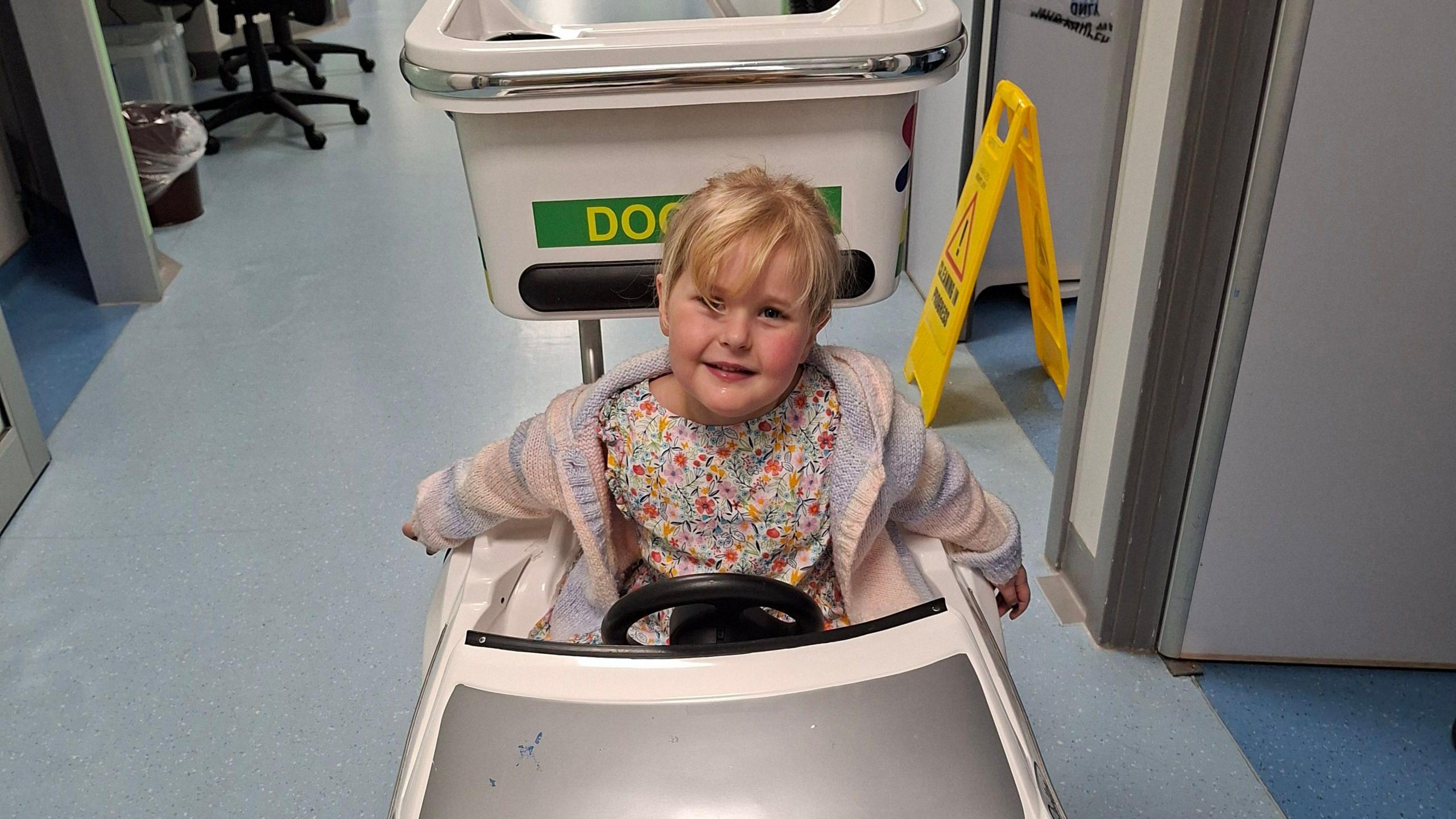 A five-year-old girl with blonde hair is sitting in a child's toy ambulance. It appears to be in a hospital corridor. There is a yellow safety sign in the background and swivel chairs.