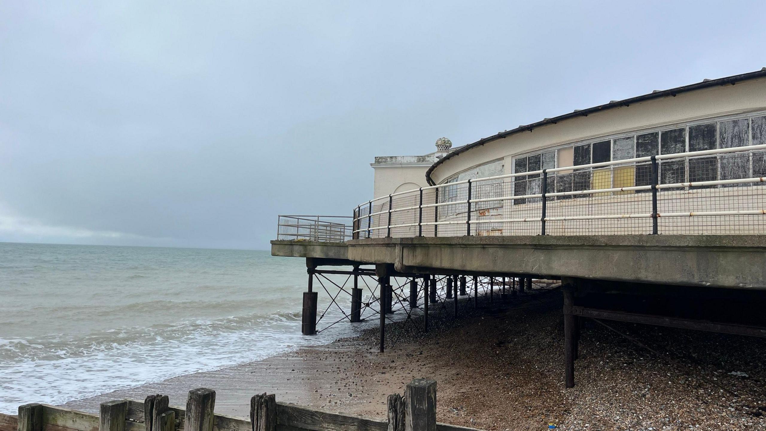 Worthing Lido which is a cream colour disc shaped building which overhangs the sea on metal stilts. The ocean is lapping at the metal structure supporting the building which is the council say it at risk due to climate change. There is metal fencing around the edge of the building.