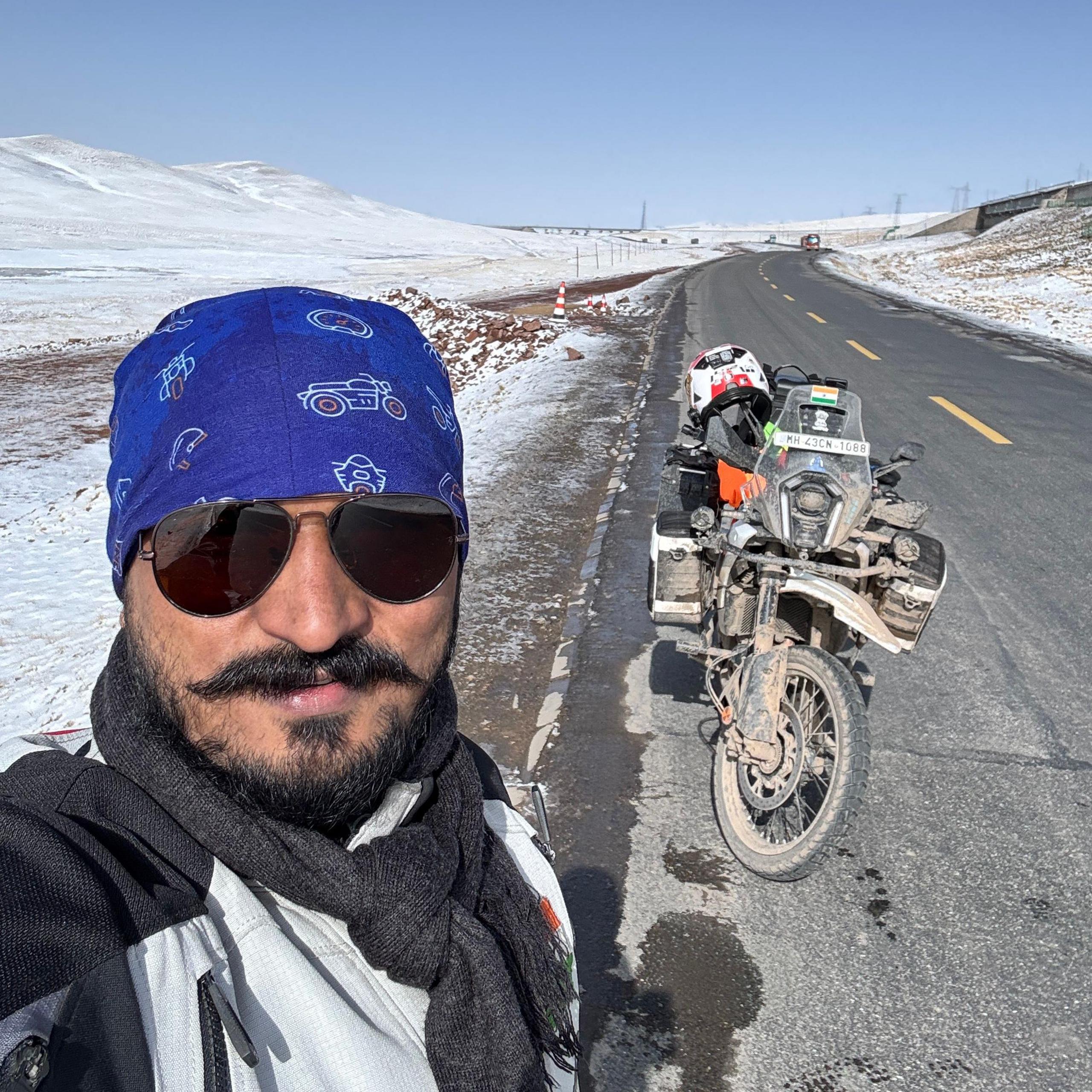 A man stands next to a motorbike wearing sunglasses and a scarf. There are snow covered hills behind him and an empty road