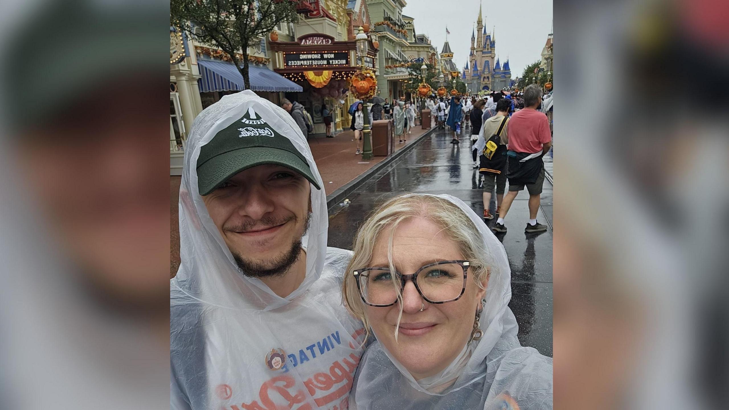 A man and a woman standing in front of the Cinderella Castle at Disney World. They are wearing clear rain ponchos