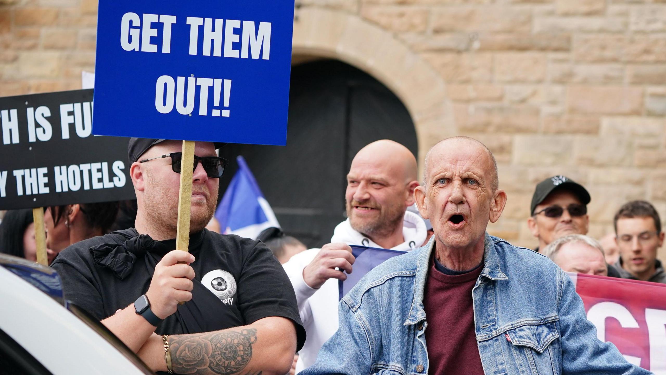 Two men at a protest - one wears a black t-shirt and sunglasses and is holding a sign which reads "get them out!!". The other man is balding, wears a denim jacket and purple t-shirt, and appears to be shouting