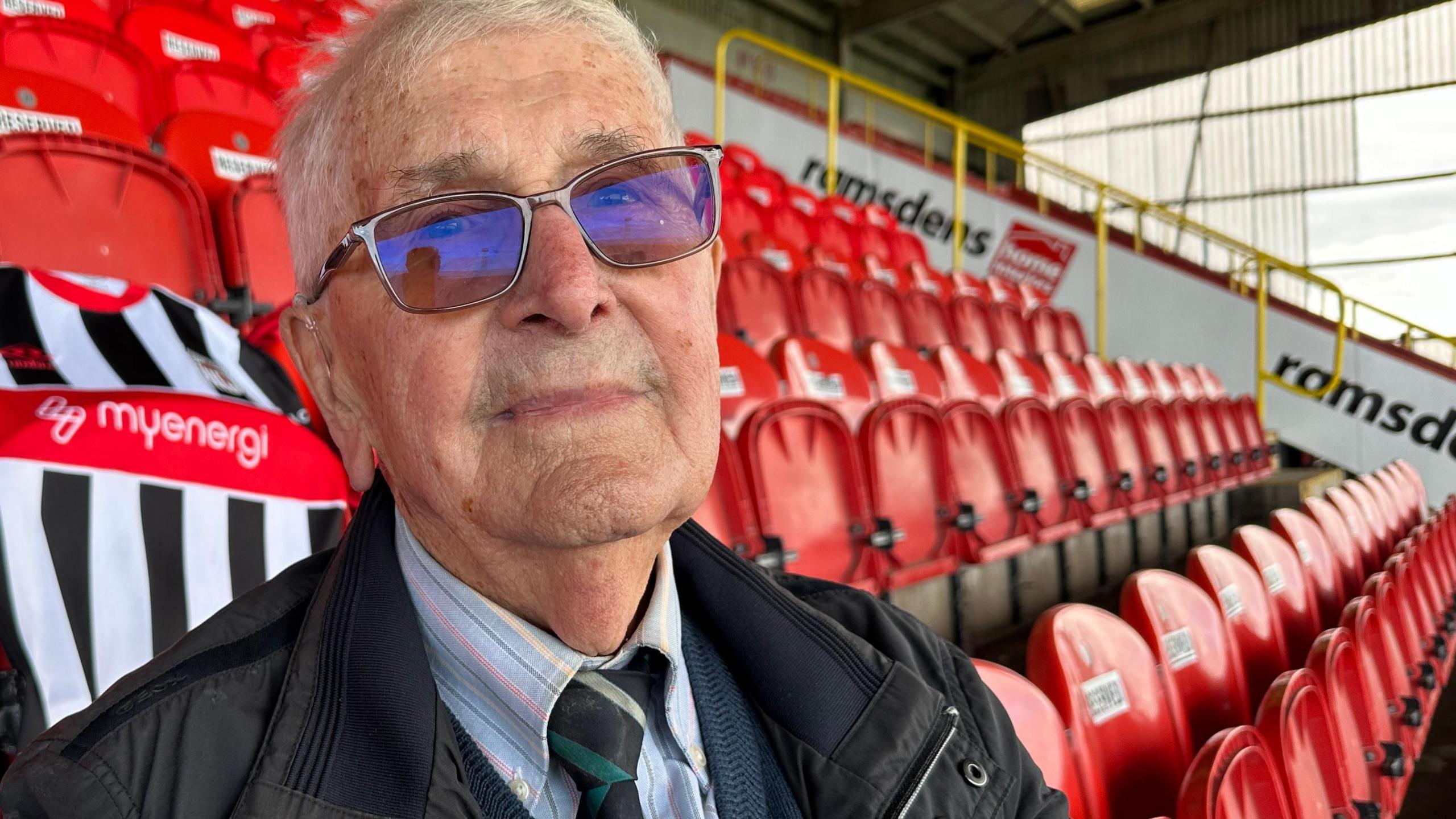 A man with short hair wearing glasses, dark jacket, a shirt and tie. He is sitting in a football stand in front of rows of folded red seats. Behind him is a black and white striped Grimsby Town shirt. On the shirt is the logo of the club sponsor, Myenergi, written in white letters on a red background.