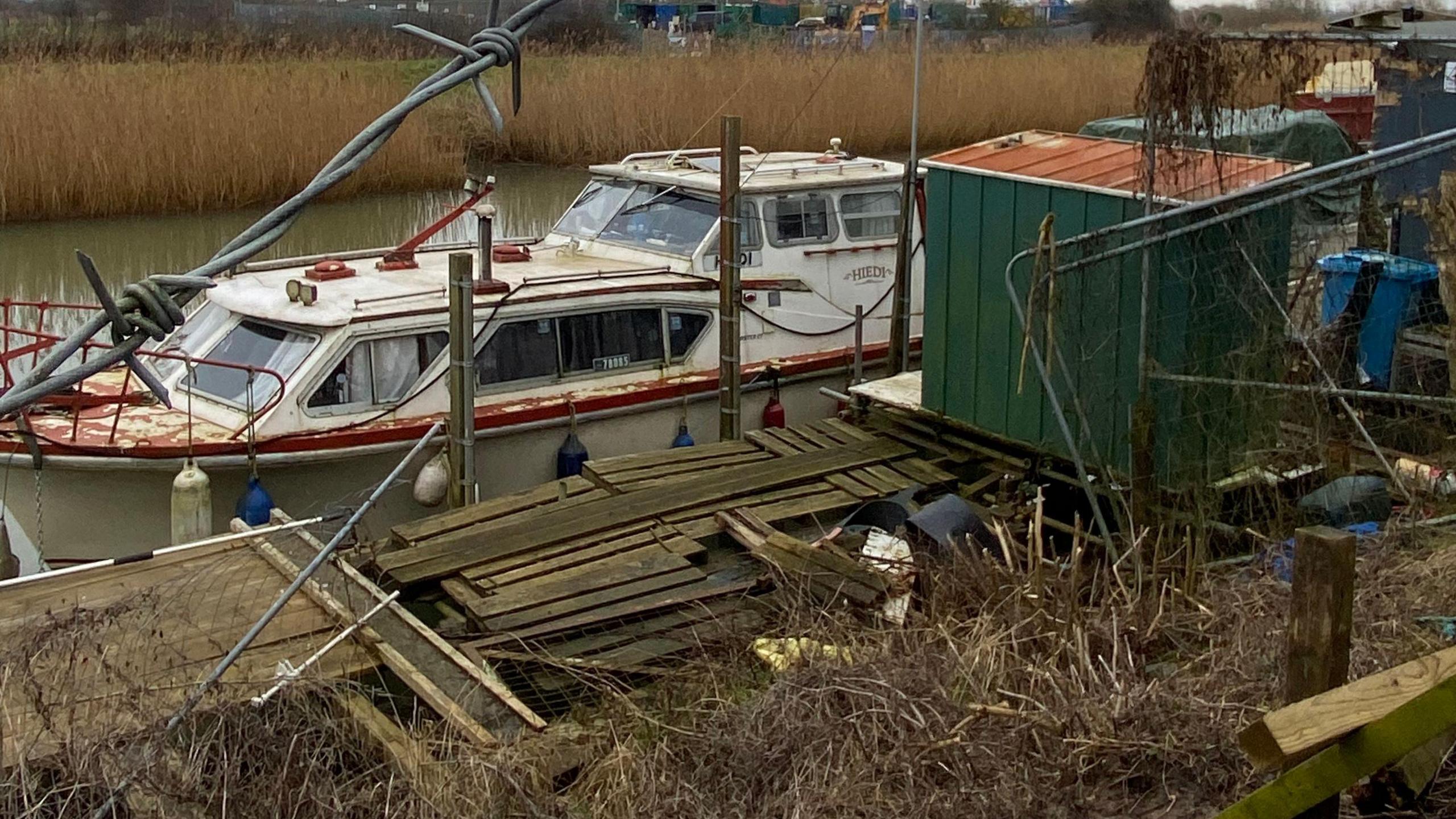 A boat that looks abandoned on the River Hull, with broken wooden scaffolding surrounding it and broken fencing